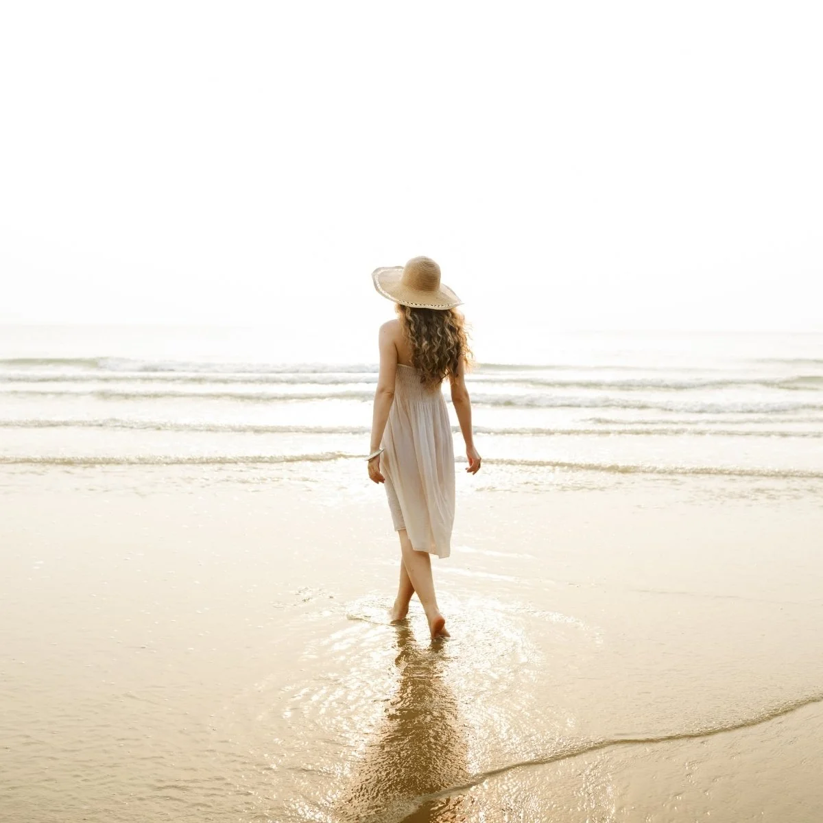 A woman with long curly hair wearing a beige dress and a wide-brimmed straw hat walking barefoot on the sand along the shoreline of a beach with gentle waves.