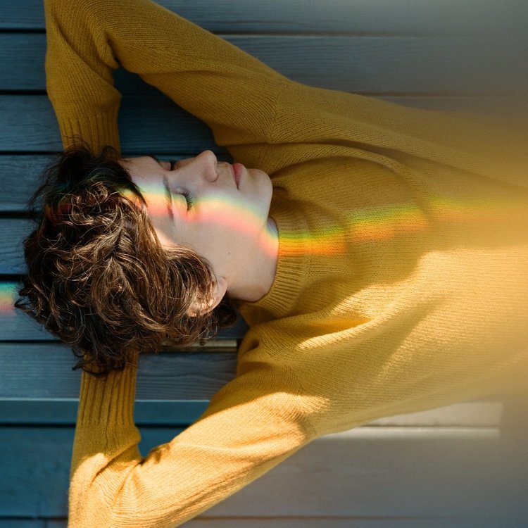 A person with curly hair lying on a wooden surface, wearing a yellow sweater, with sunlight casting a rainbow-colored light across their face.