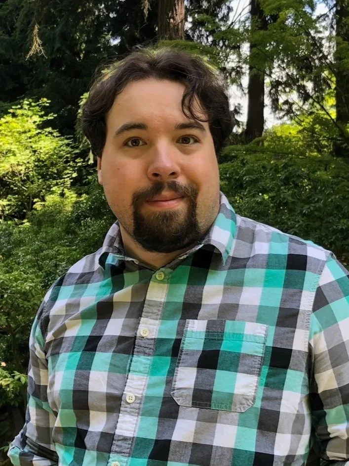 A man with dark hair and a beard posing outdoors in front of green trees, wearing a plaid shirt.