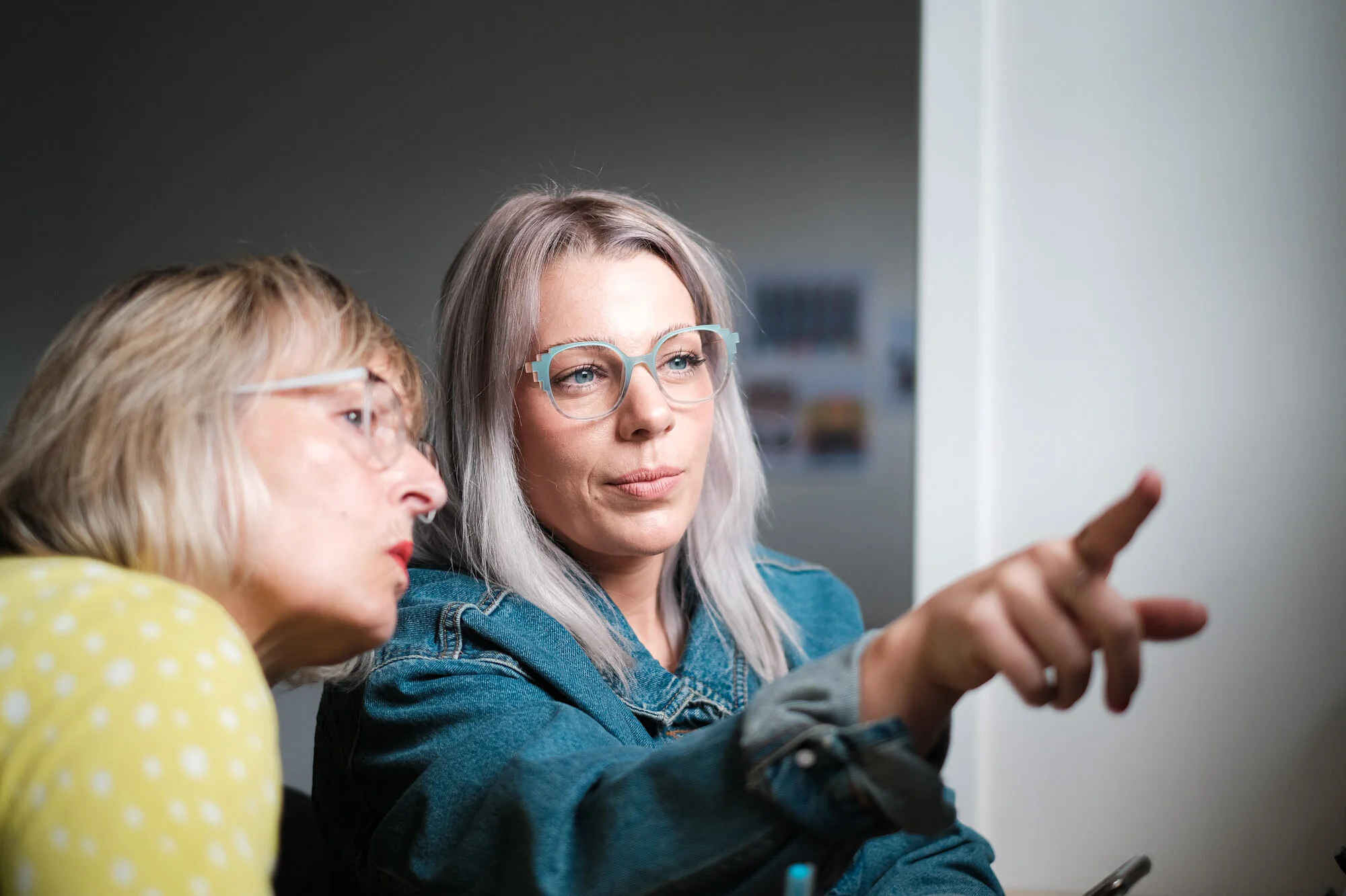 Two women wearing glasses, one in a yellow top and the other in a denim jacket, looking and pointing at something out of view.