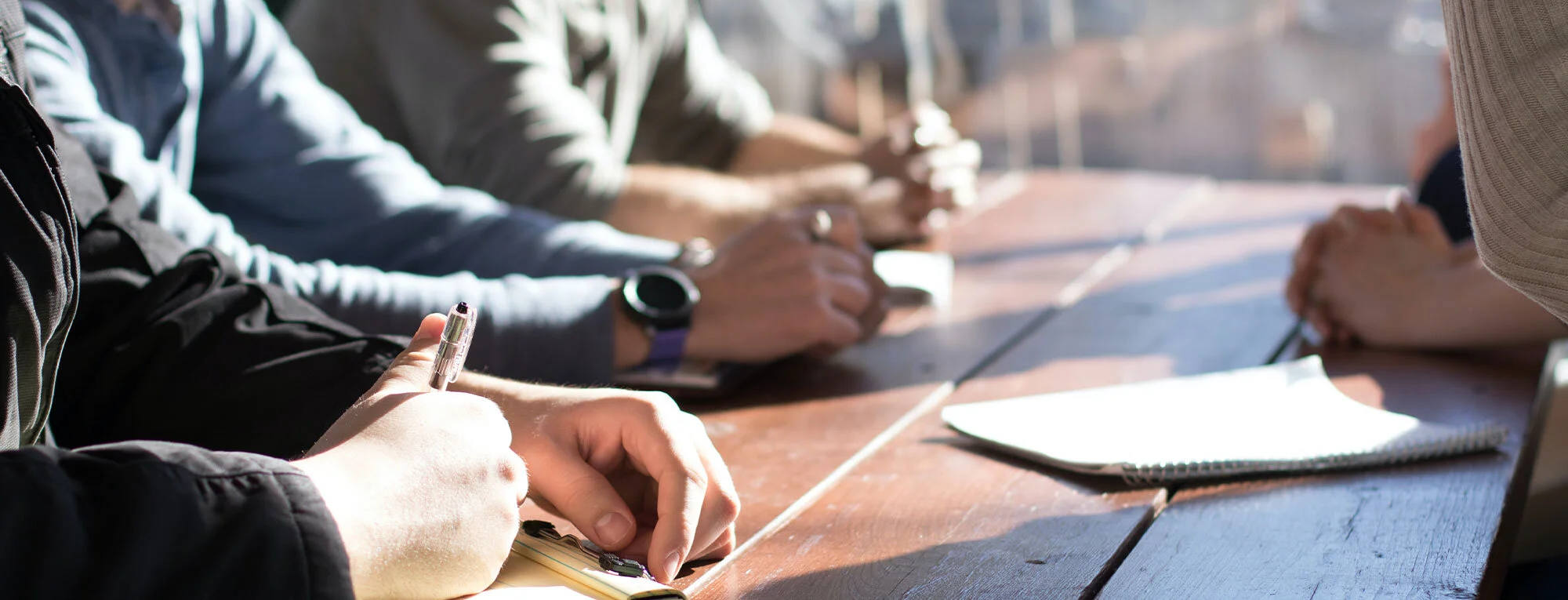 Group of people sitting around a wooden table with notebooks and pens, engaged in a meeting or discussion.
