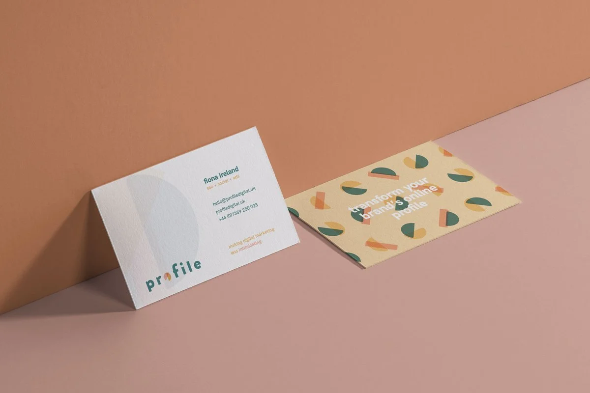 Two business cards on a pink surface, featuring text and geometric patterns. One card includes contact details with a minimalist design, and the other has the phrase "transform your brand’s online profile" with abstract shapes.