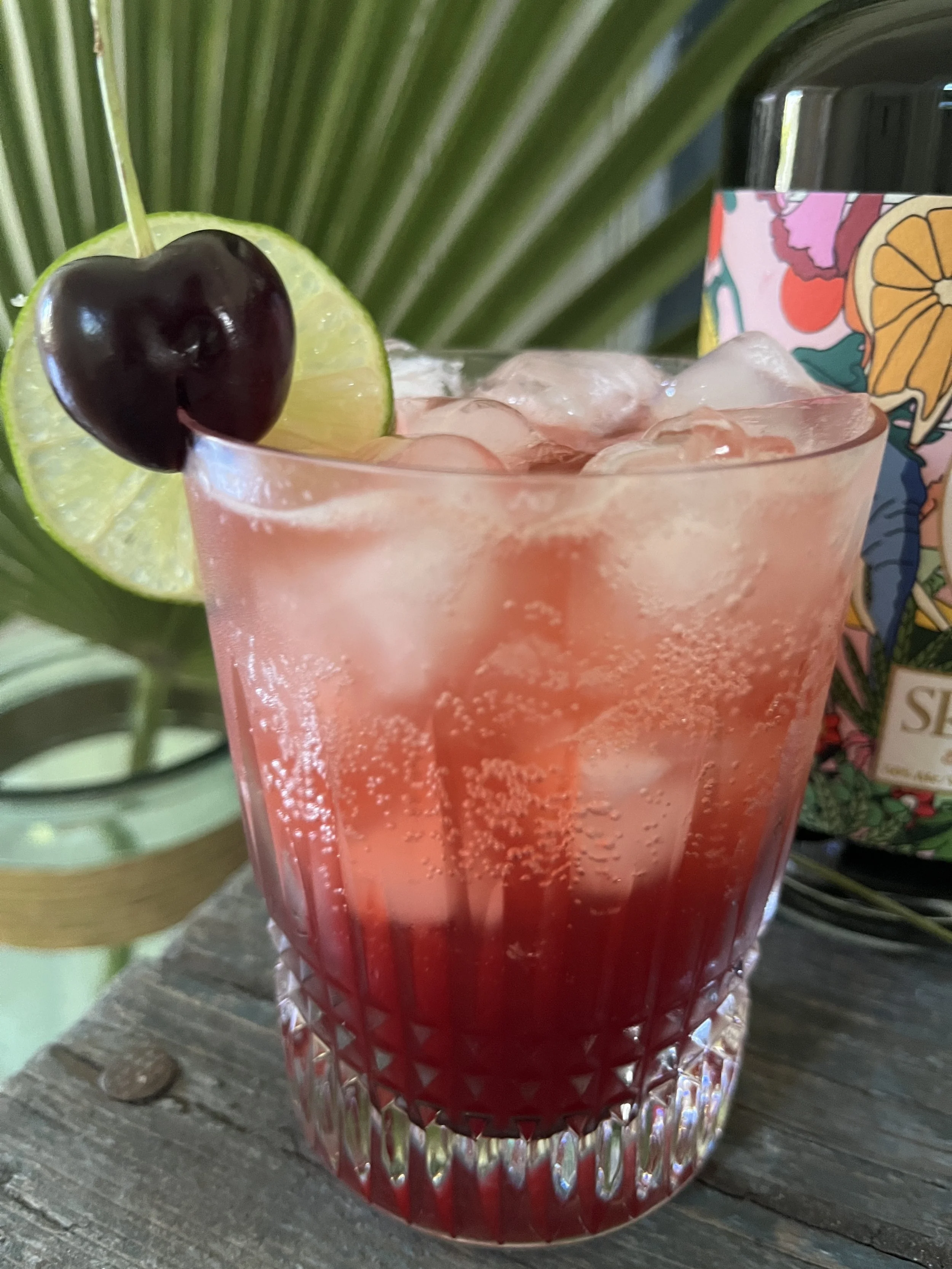 Elephant Rickey is adapted from Brad Thomas Parsons cocktail Ramazotti Rickey. Cocktail with ice in a textured glass, garnished with a lime slice and a cherry, placed on a wooden surface with a tropical leaf and a patterned bottle in the background.