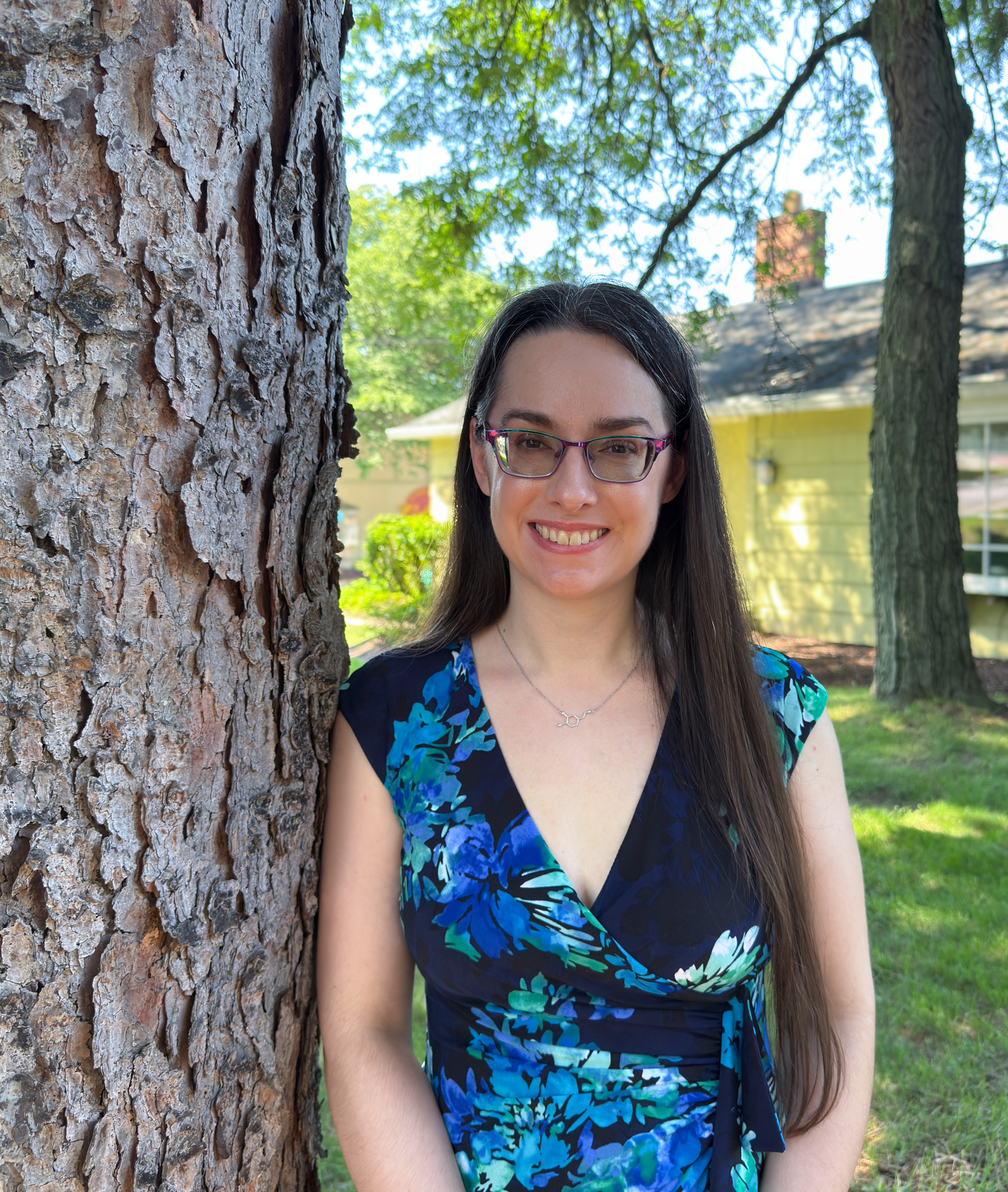 Woman standing in a blue dress next to a tree in front of a yellow house wearing glasses
