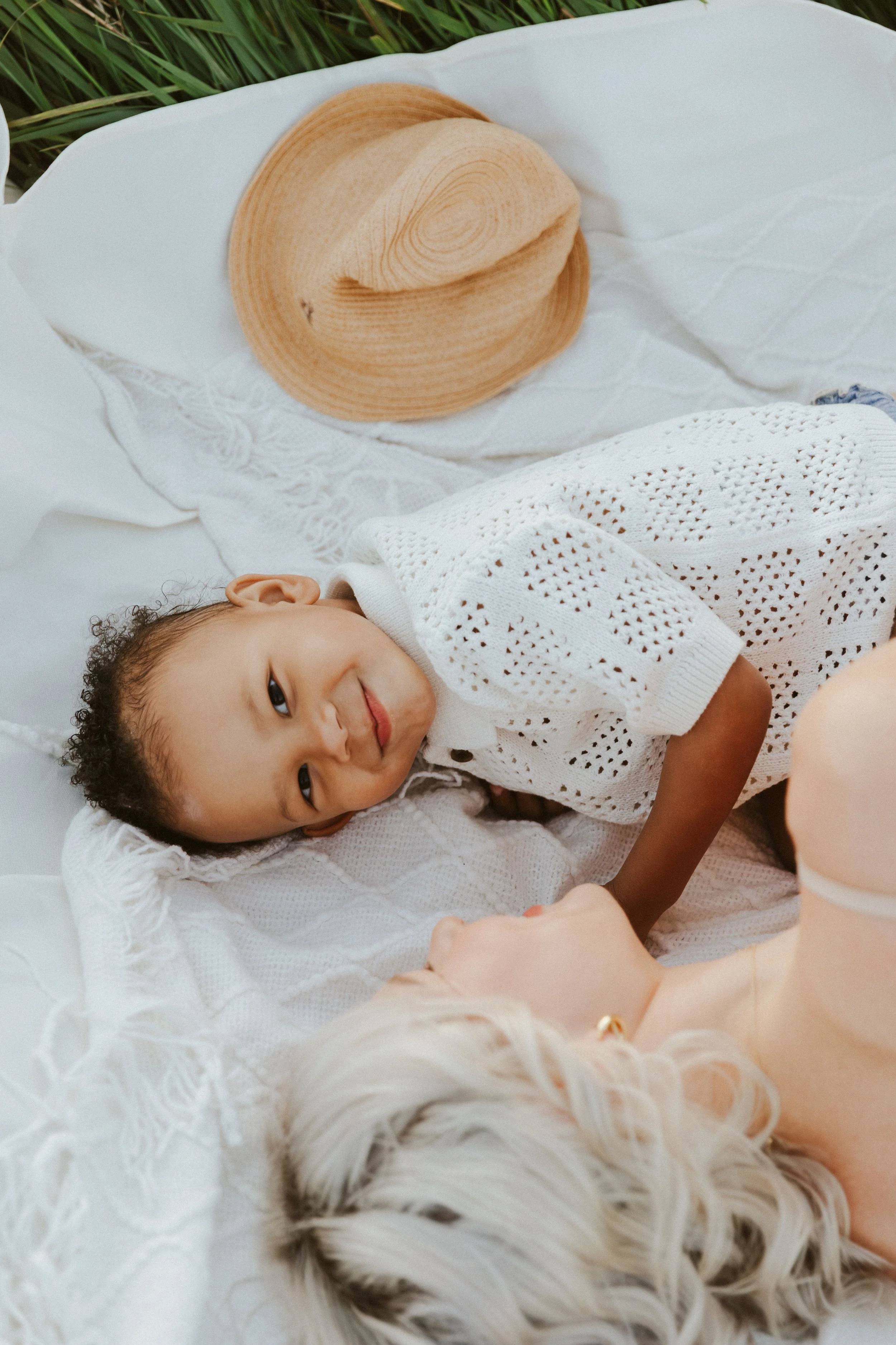 A young child lying on a white textured blanket outdoors, smiling and holding hands with an adult with short, wavy blonde hair.