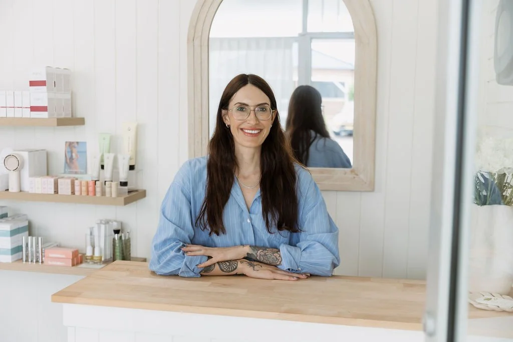 A woman with long brown hair, wearing glasses and a blue shirt, sits behind a wooden counter in a bright, white-walled shop with skincare products on shelves, smiling at the camera.