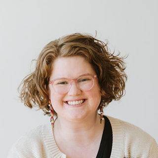 A woman with curly hair, wearing glasses, earrings, a beige sweater, and a black top, smiling against a light background.