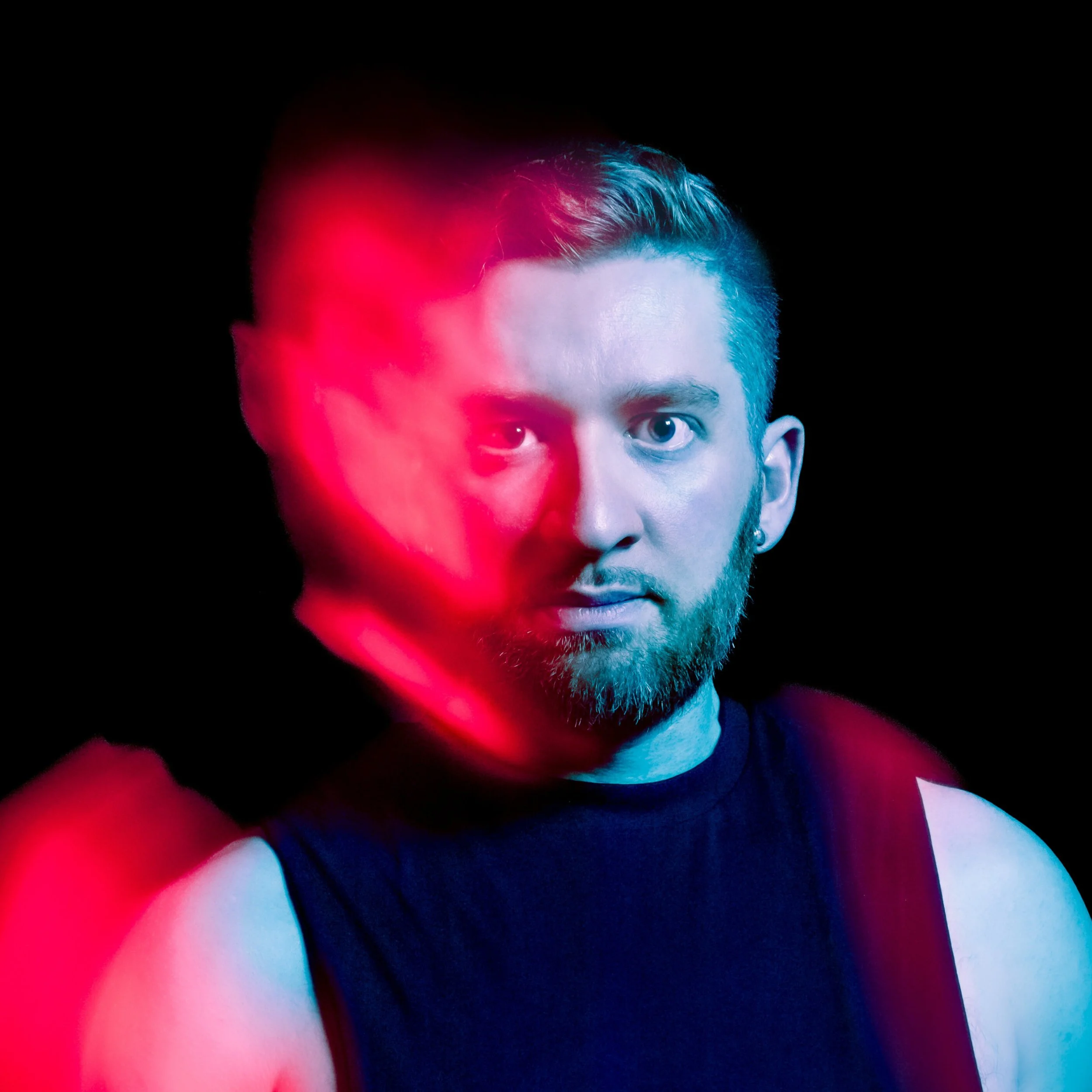 A portrait of a young man with short hair, a beard, and blue eyes, wearing a black sleeveless shirt, illuminated with blue and pink lighting against a black background.