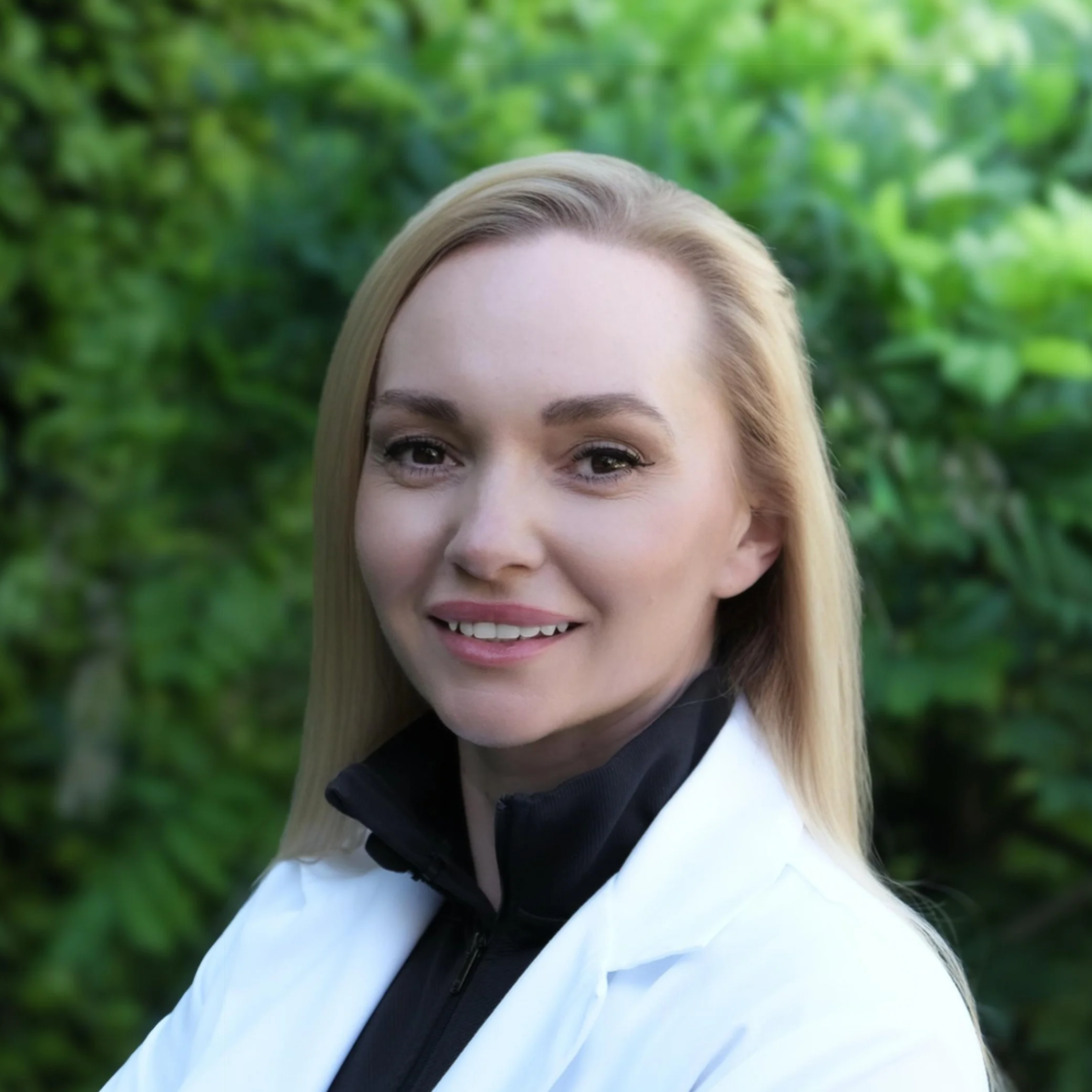 Close-up of a smiling woman with blonde hair and light skin, wearing a white jacket with a black collar, outdoors with green foliage in the background.