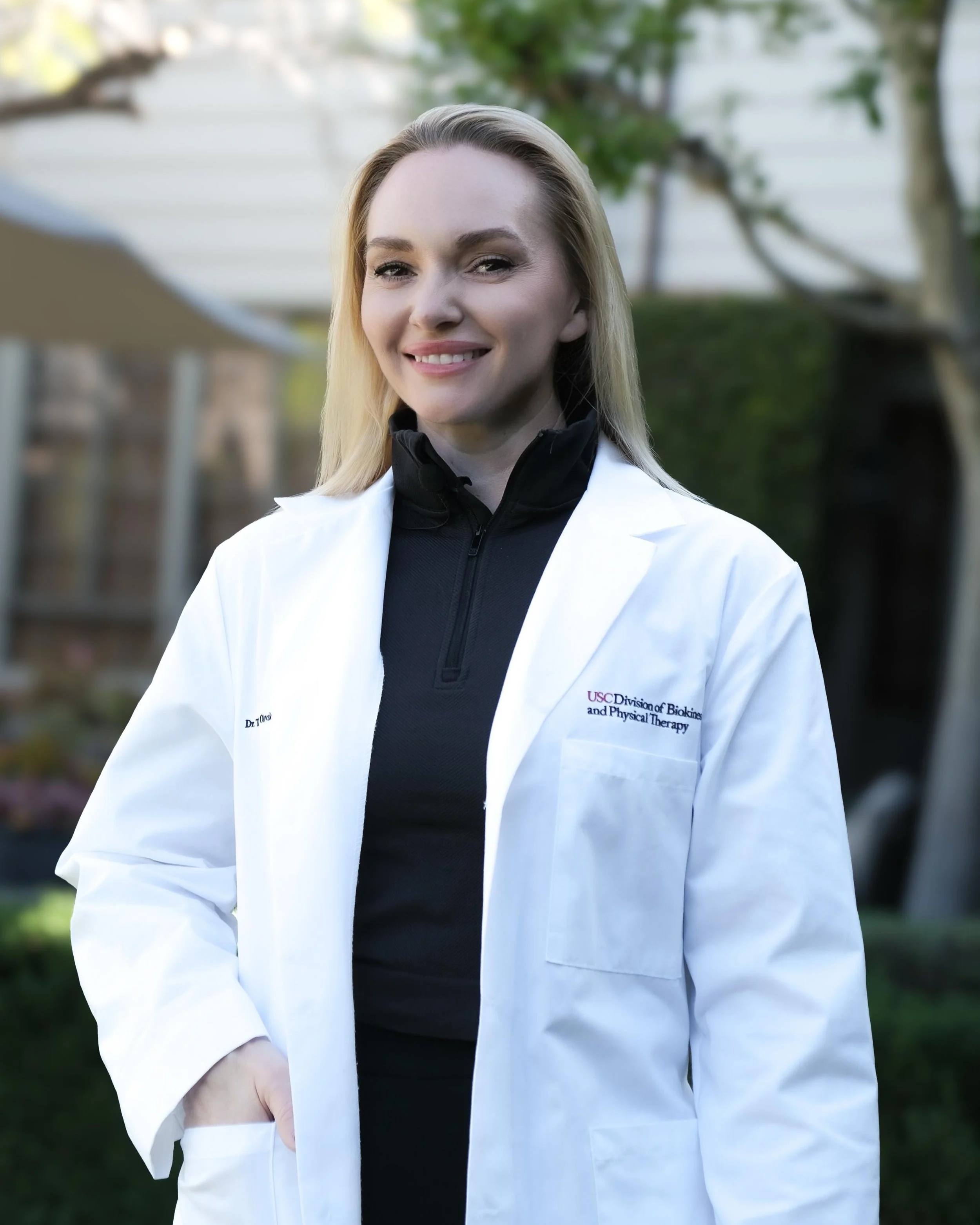 Smiling woman in a white lab coat and black athletic top standing outdoors with green trees and white house in the background.