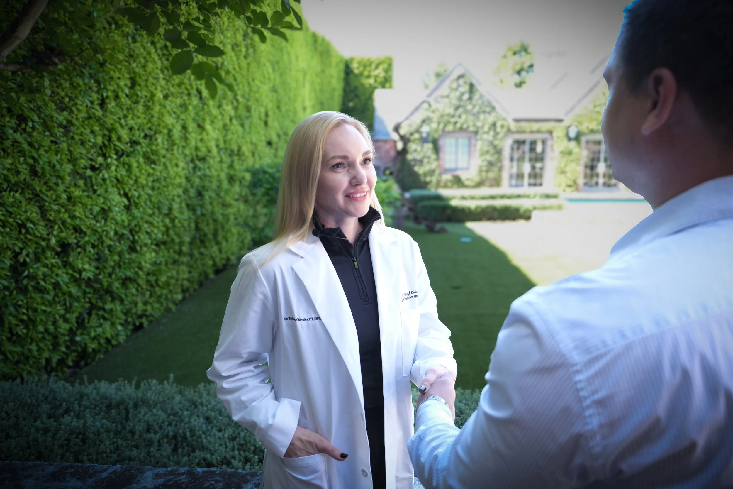 A woman in medical attire shaking hands with a man outdoors, with a large house and greenery in the background.