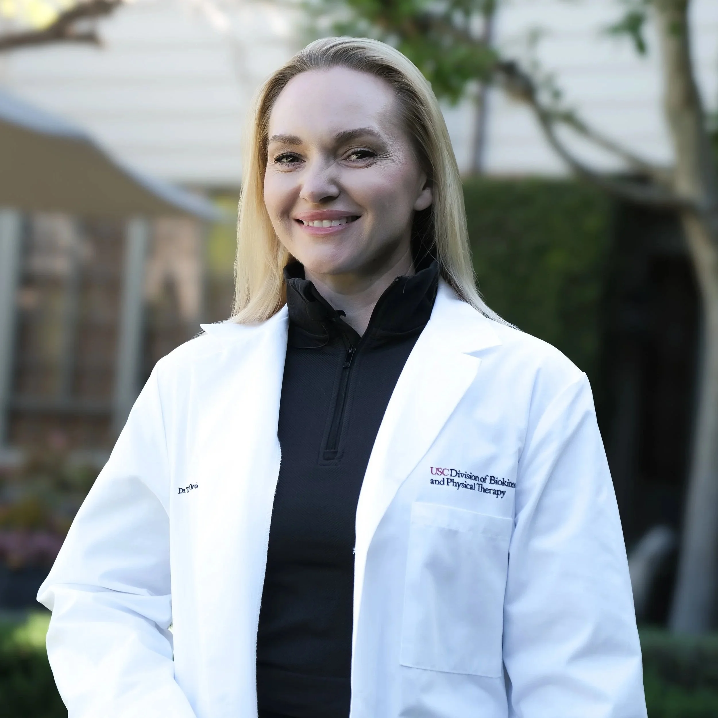 A woman in a white lab coat smiling outdoors, with trees and blurred background.