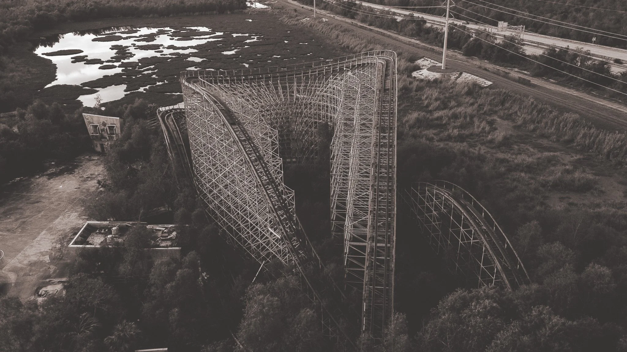 An aerial view of an abandoned wooden roller coaster surrounded by trees, with wetlands and a highway in the background.