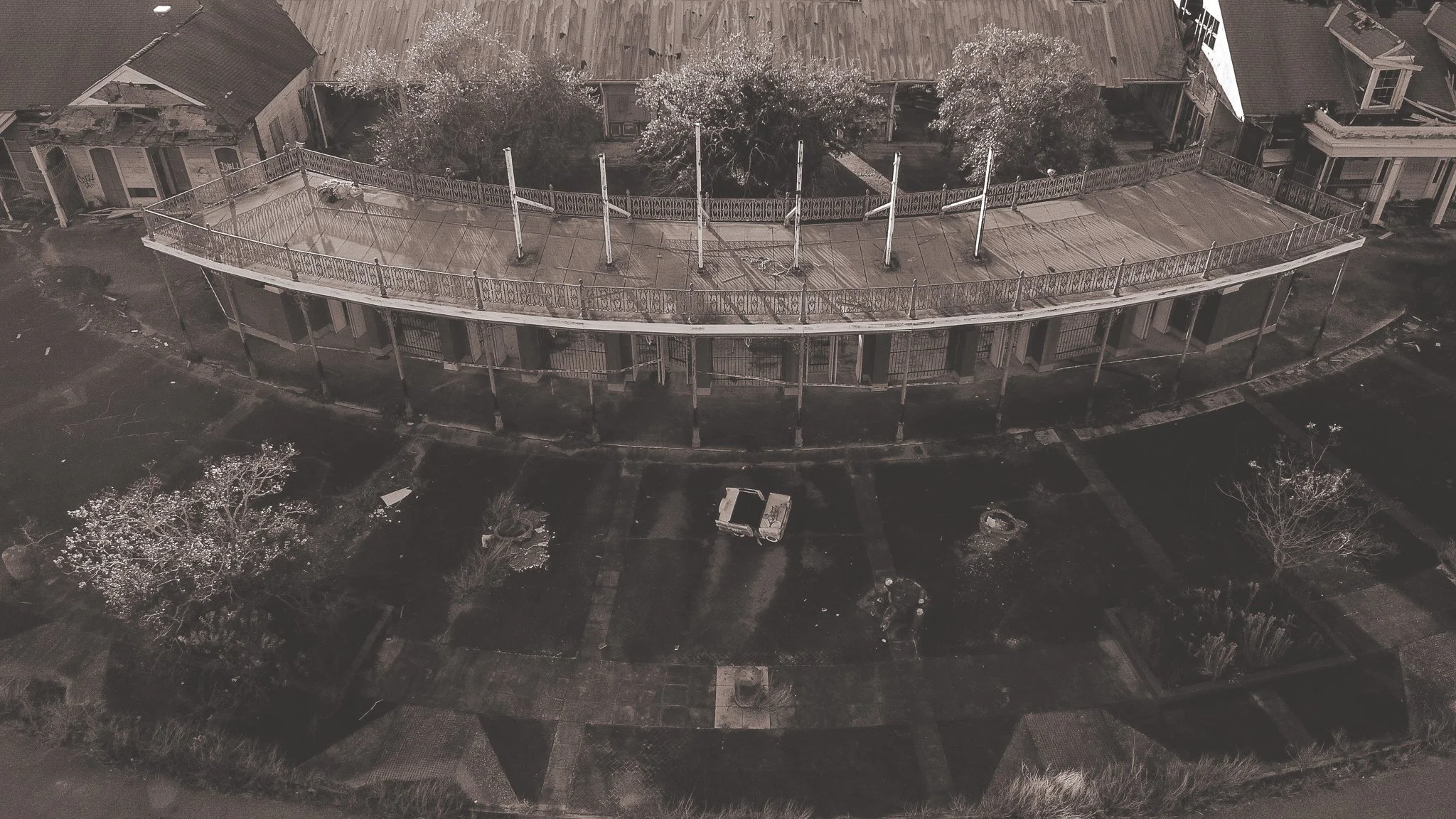 An aerial drone view of a covered porch with a decorative railing, lower level houses, a small garden with trees, and a paved pathway in front. the abandoned Six Flags New Orleans Jazzland. This iconic, world-famous park was devastated 
