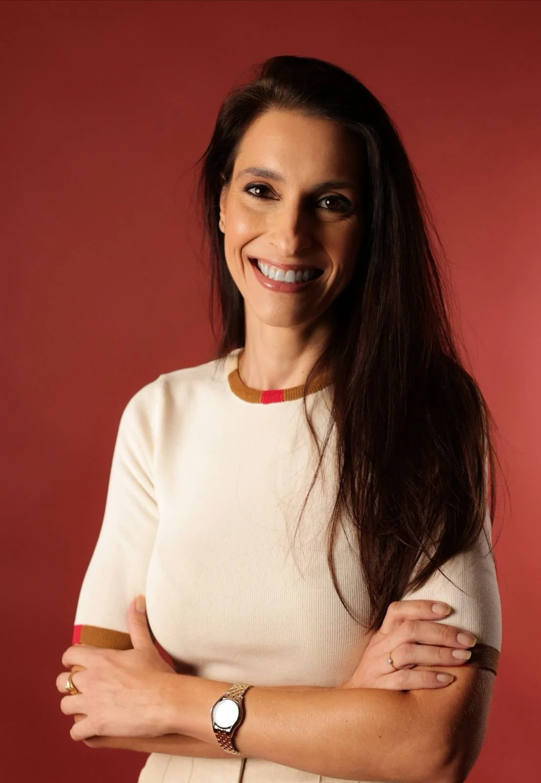 Mulher sorridente com pele clara, cabelo castanho escuro, usando uma blusa de cor creme com detalhes em marrom e vermelho no pescoço e nas mangas, em pé contra fundo vermelho