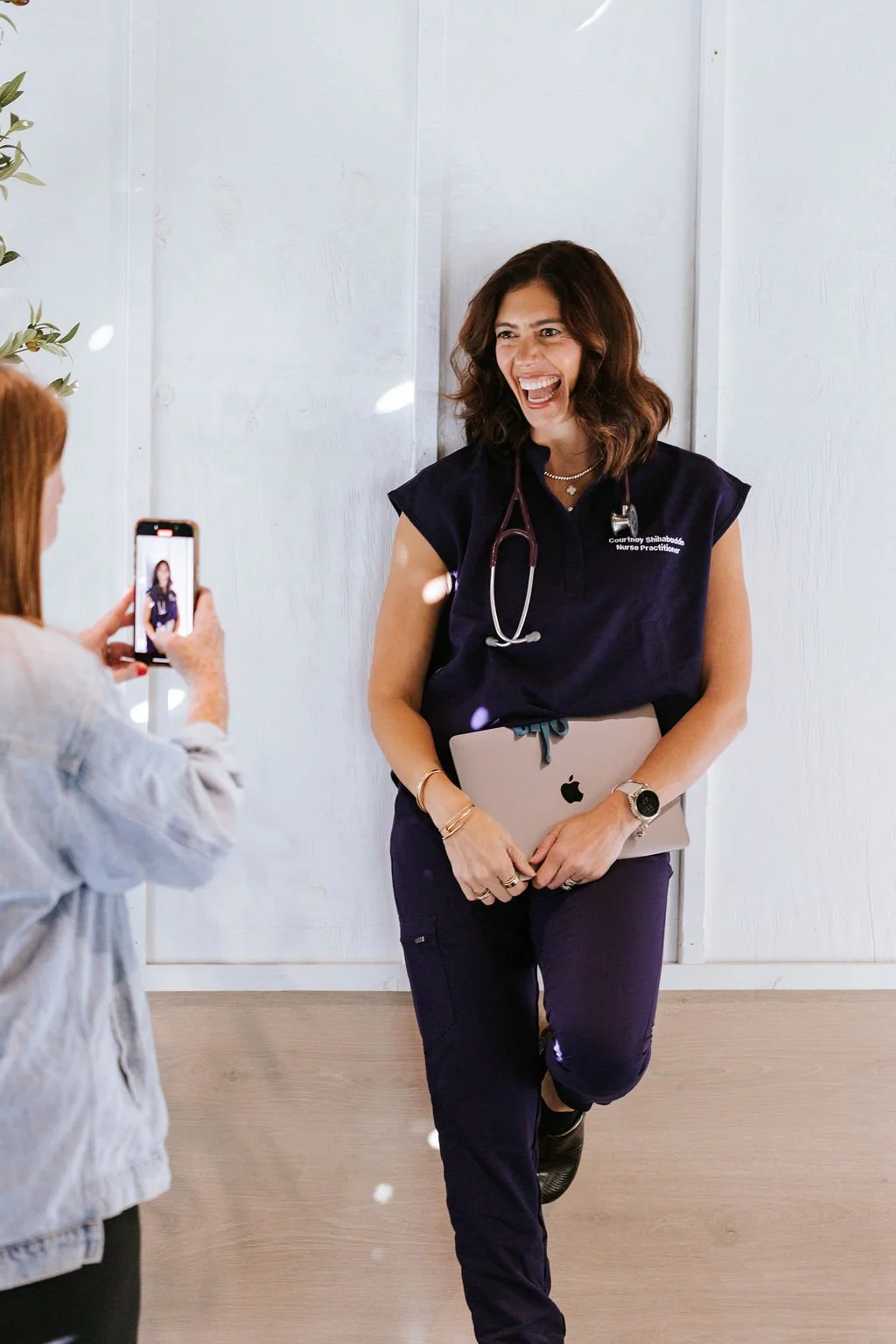 Female nurse practitioner with stethoscope around neck, holding a laptop, smiling and standing against a white wall while being photographed by a woman with red hair.