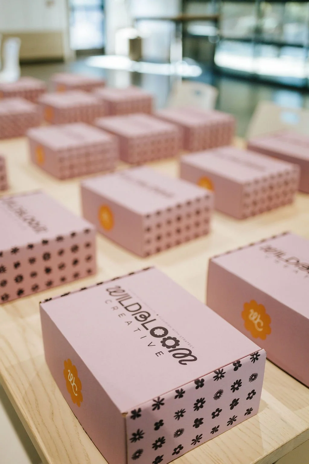 Pink boxes with black floral patterns and yellow logos, arranged on a wooden table in a well-lit room with large windows.
