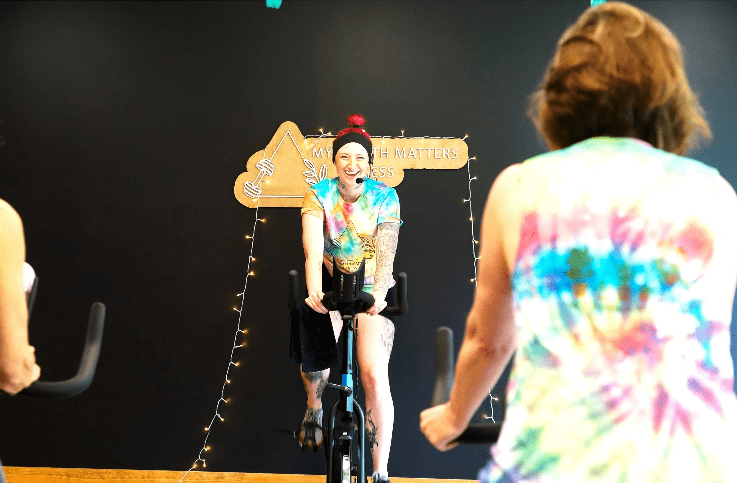 A woman with a microphone headset riding a stationary bike in a fitness class, smiling, wearing a colorful tie-dye shirt, with guest instructors in tie-dye shirts participating. The background has string lights and a wooden sign that reads 'My health