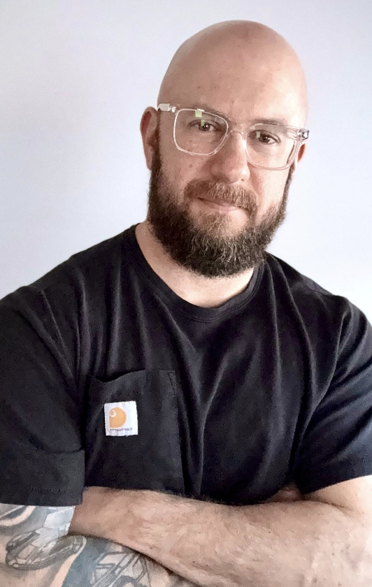 A bald man with glasses and a beard, wearing a black t-shirt, standing against a plain light-colored background.
