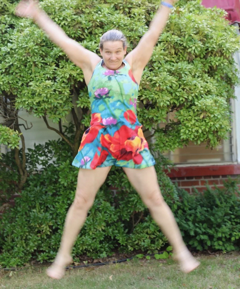 A woman jumping in front of a green bush wearing a colorful floral romper.