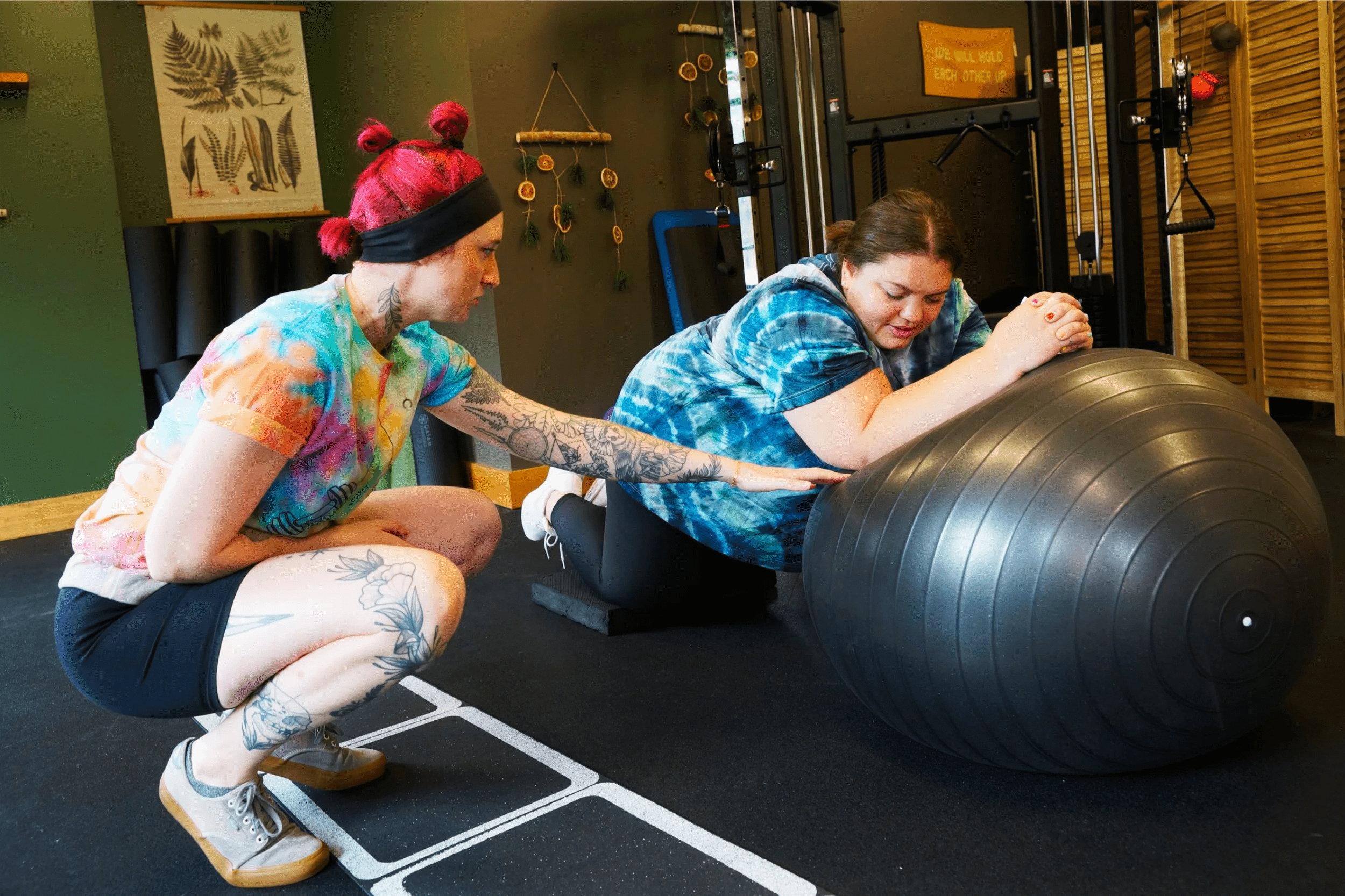 A woman with tattoos and pink hair helping another woman in a tie-dye shirt exercise with a large black stability ball in a gym.