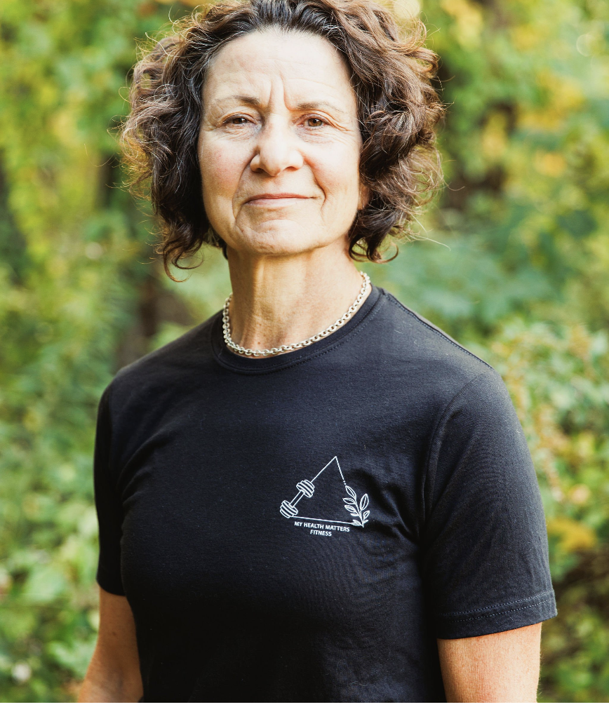 A middle-aged woman with curly brown hair outdoors, wearing a black t-shirt with a fishing hook and leaf design and the text "My Health Matters Fitness," and a pearl necklace.