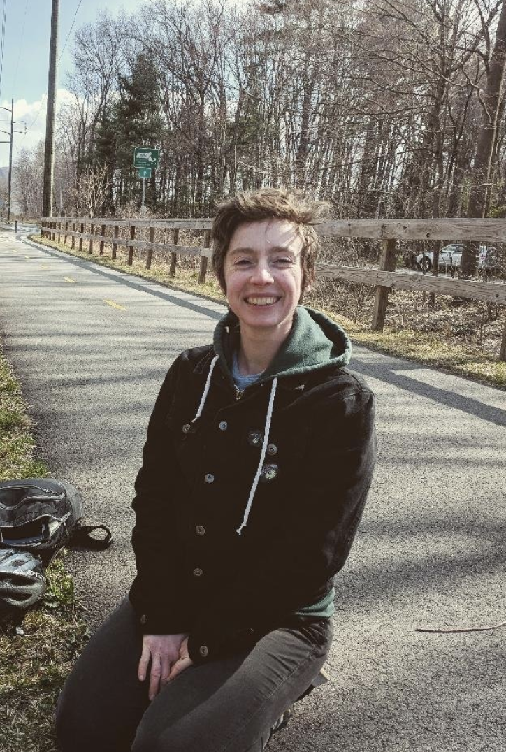 A smiling young person with short, tousled hair, wearing a black jacket, gray pants, and a green hoodie, sitting outdoors on a bike seat next to a paved bike trail with trees and a wooden fence in the background.
