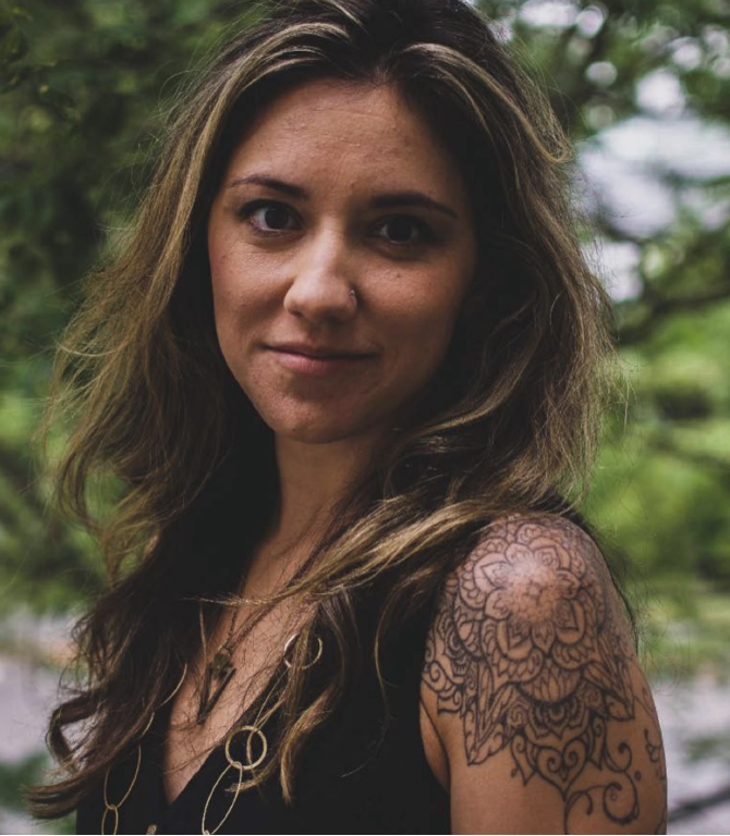 A young woman with wavy brown hair, wearing a black top and necklace, standing outdoors with greenery in the background, showing a detailed floral tattoo on her shoulder.