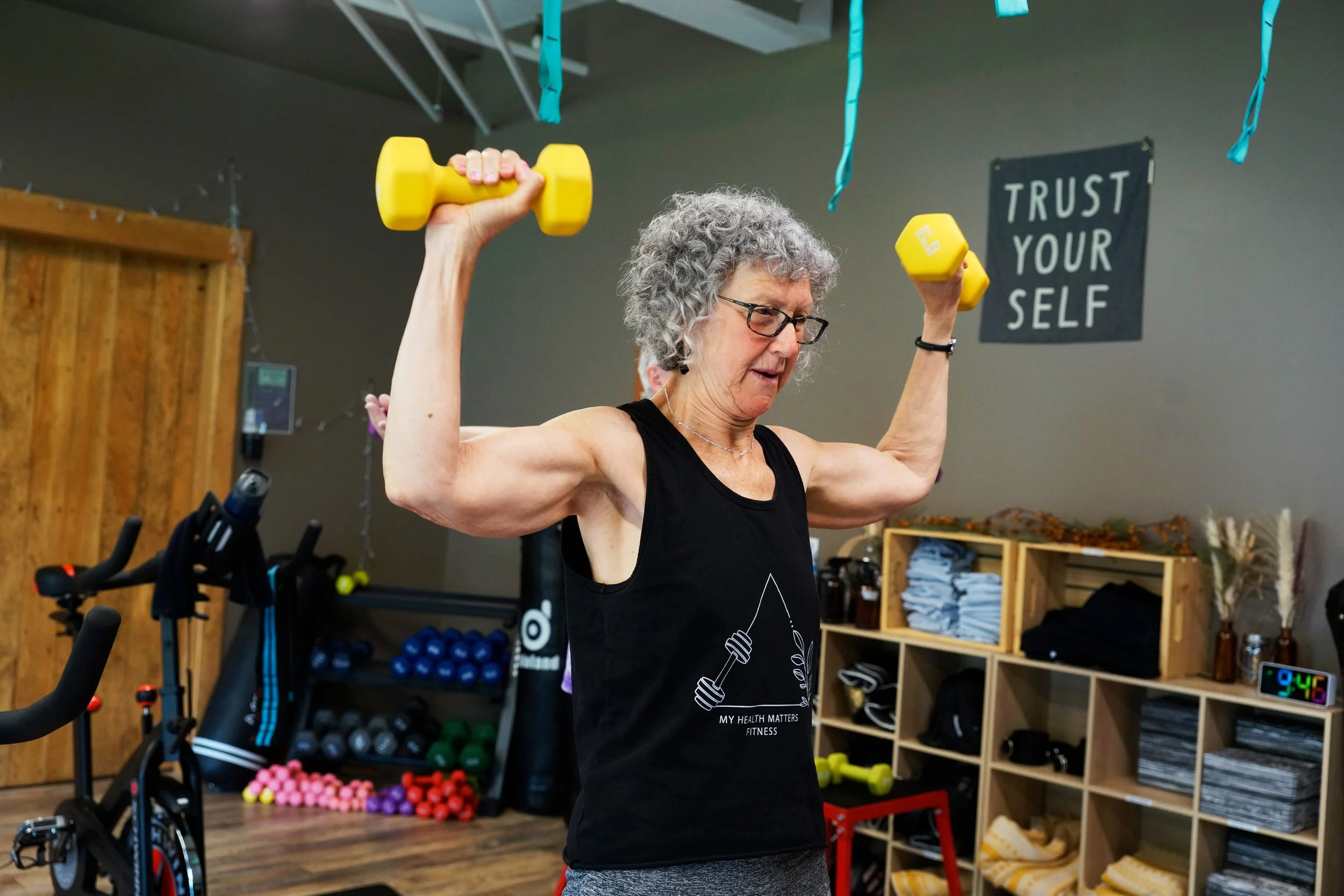An older woman with gray curly hair and glasses lifting yellow dumbbells in a fitness gym, wearing a black sleeveless shirt and gray pants.