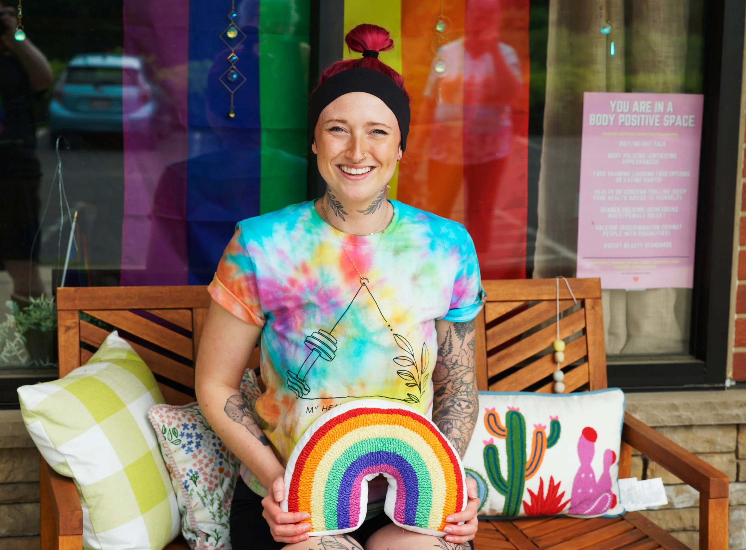 A picture of a tattoo-ed woman with pink hair sitting on a bench in front of a window that has a pride flag. She is holding a rainbow pillow and smiling.