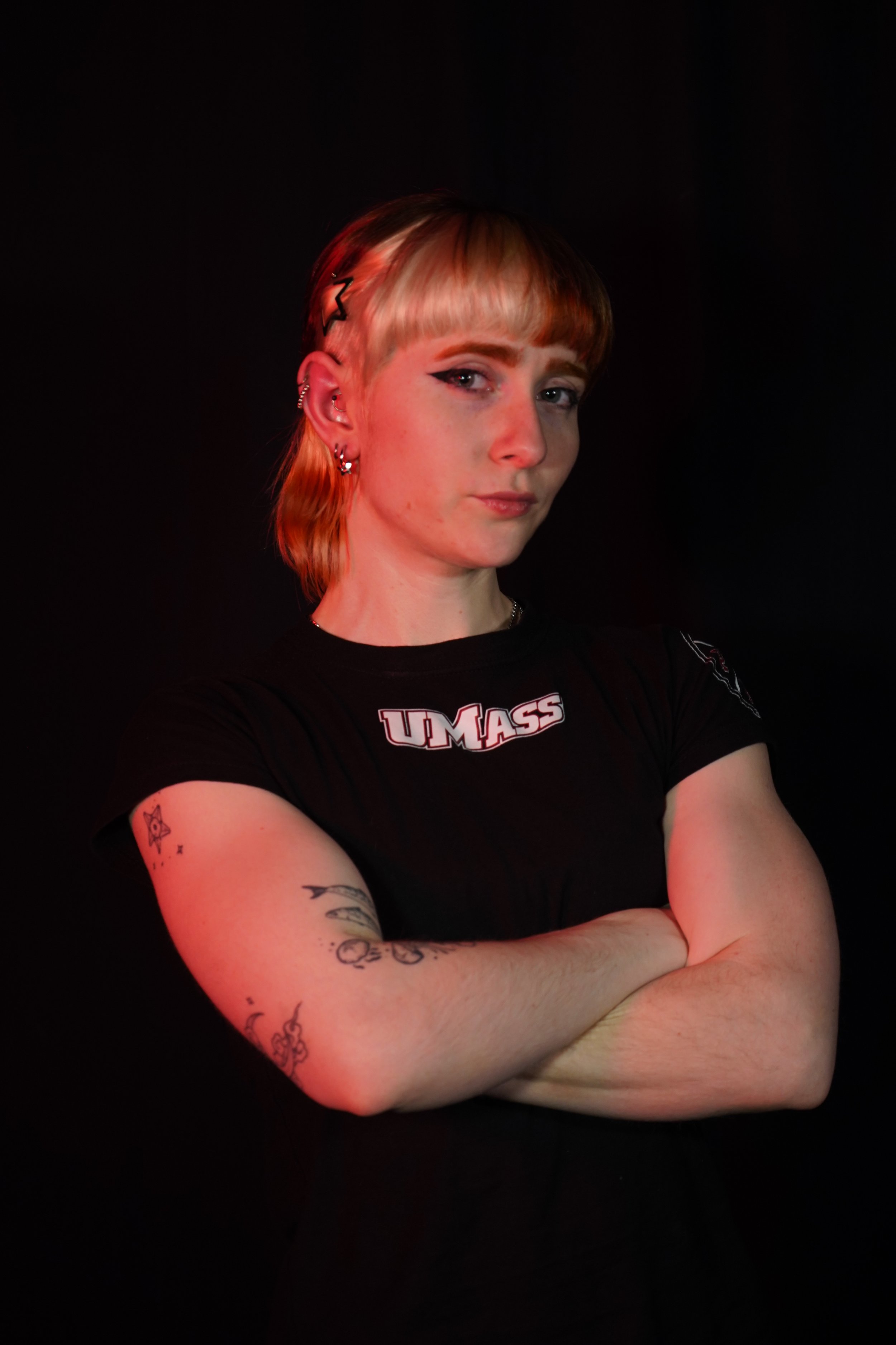 A woman with short hair wearing a black University of Massachusetts t-shirt in front of a black background