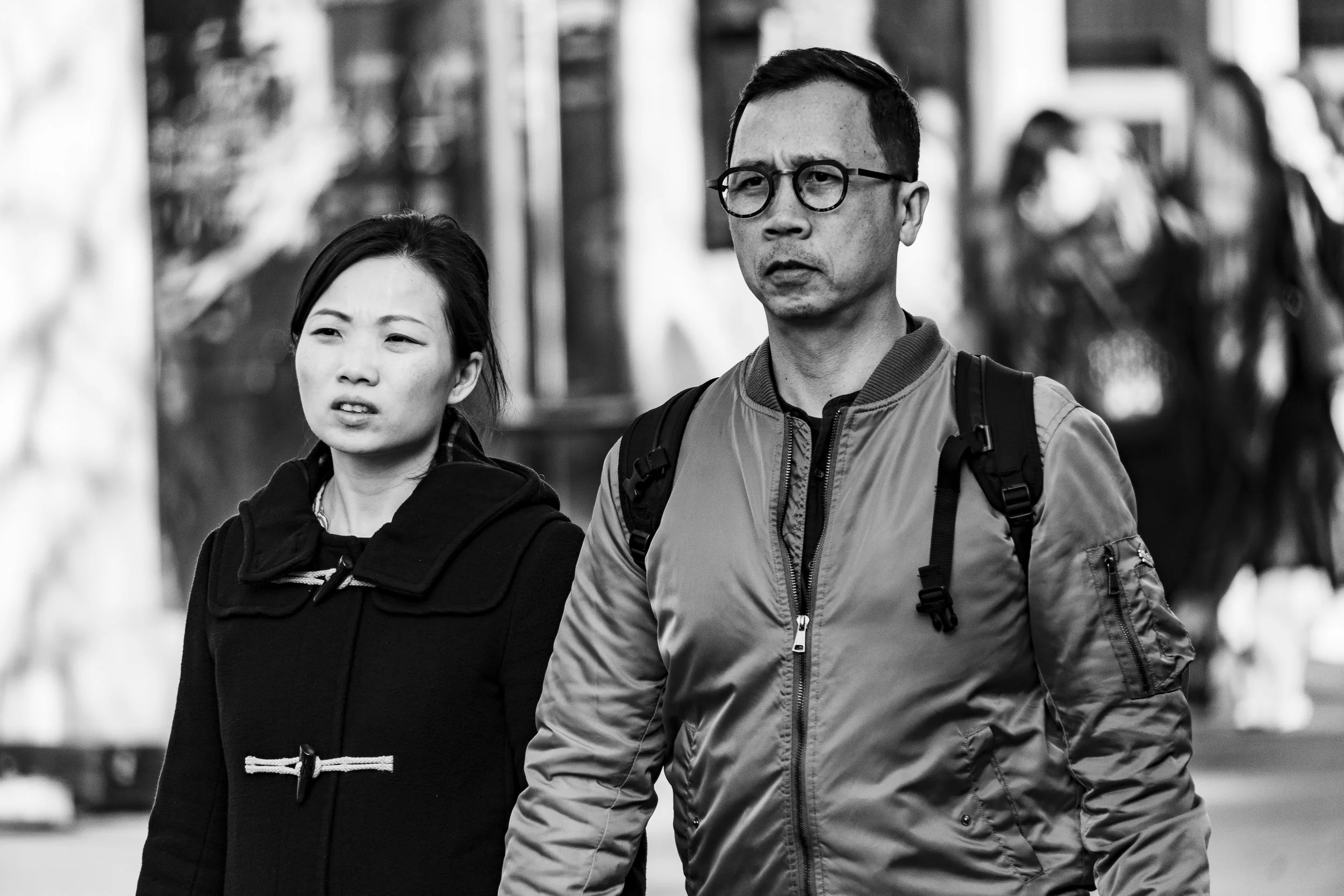 Two people walking outdoors on a city street, one woman and one man, both looking ahead with serious expressions, black and white photo