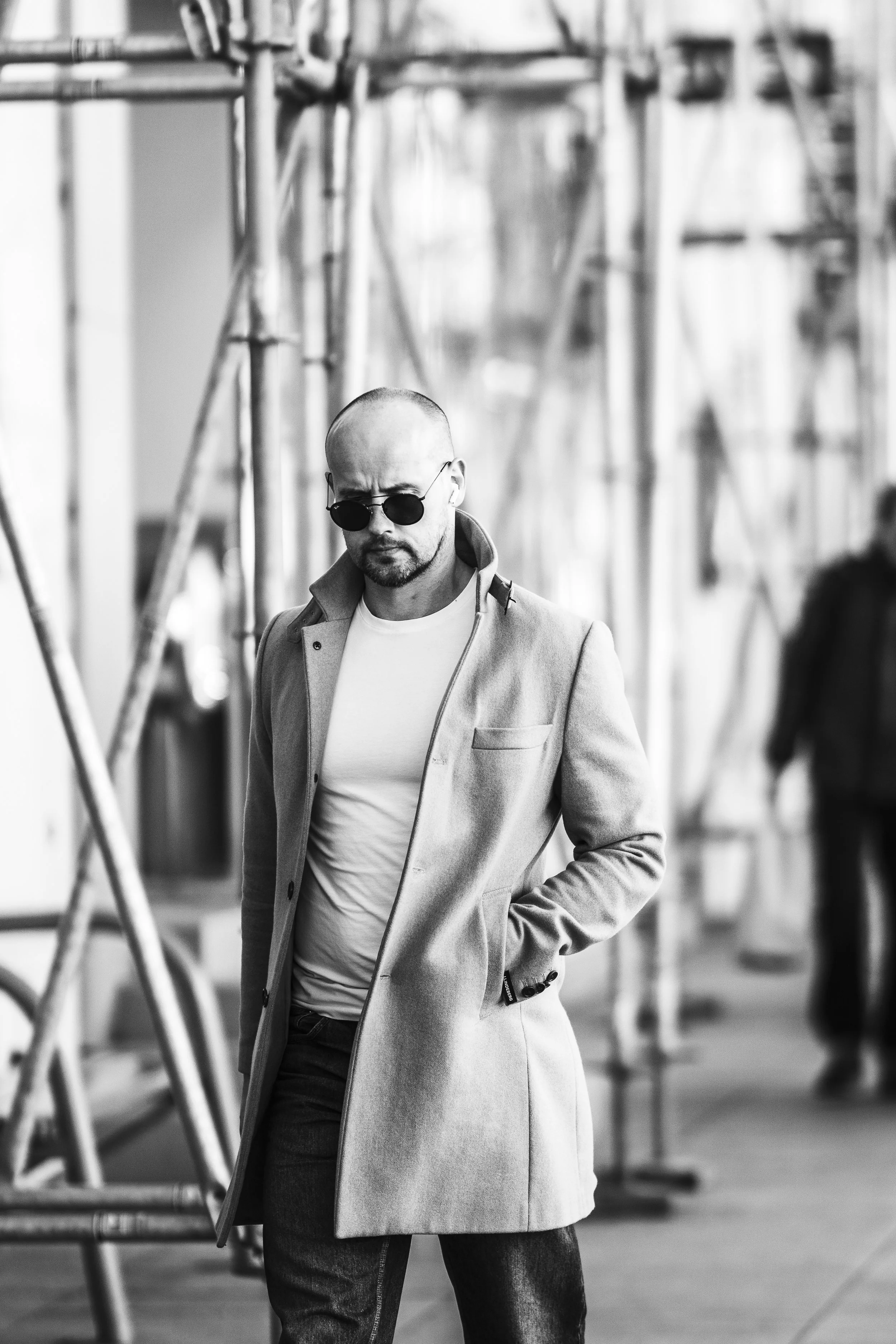 A man wearing sunglasses and a light-colored coat walking outdoors in an urban setting.