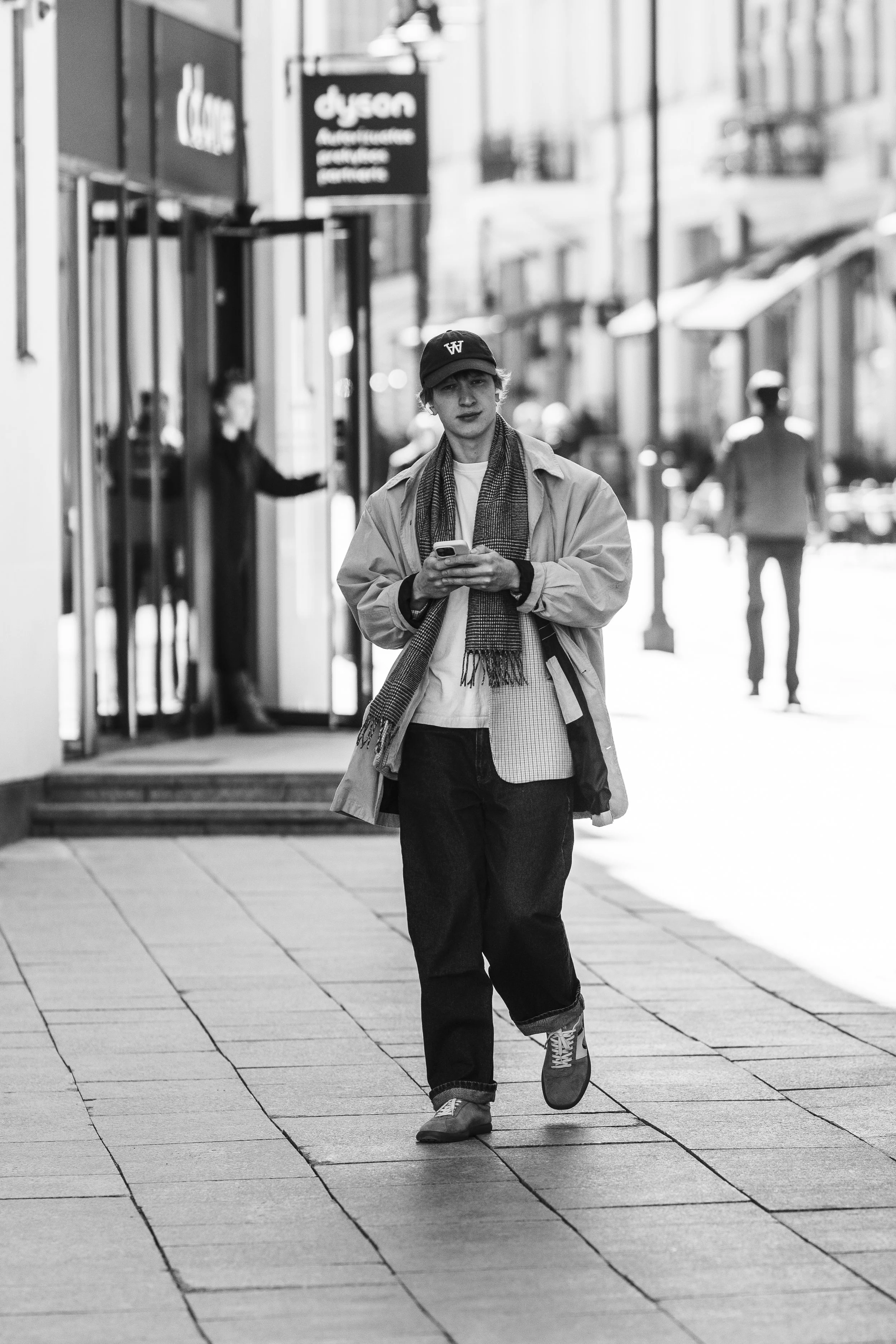 A young man walking on a city sidewalk while looking at his phone, with storefronts and pedestrians in the background, in black and white.