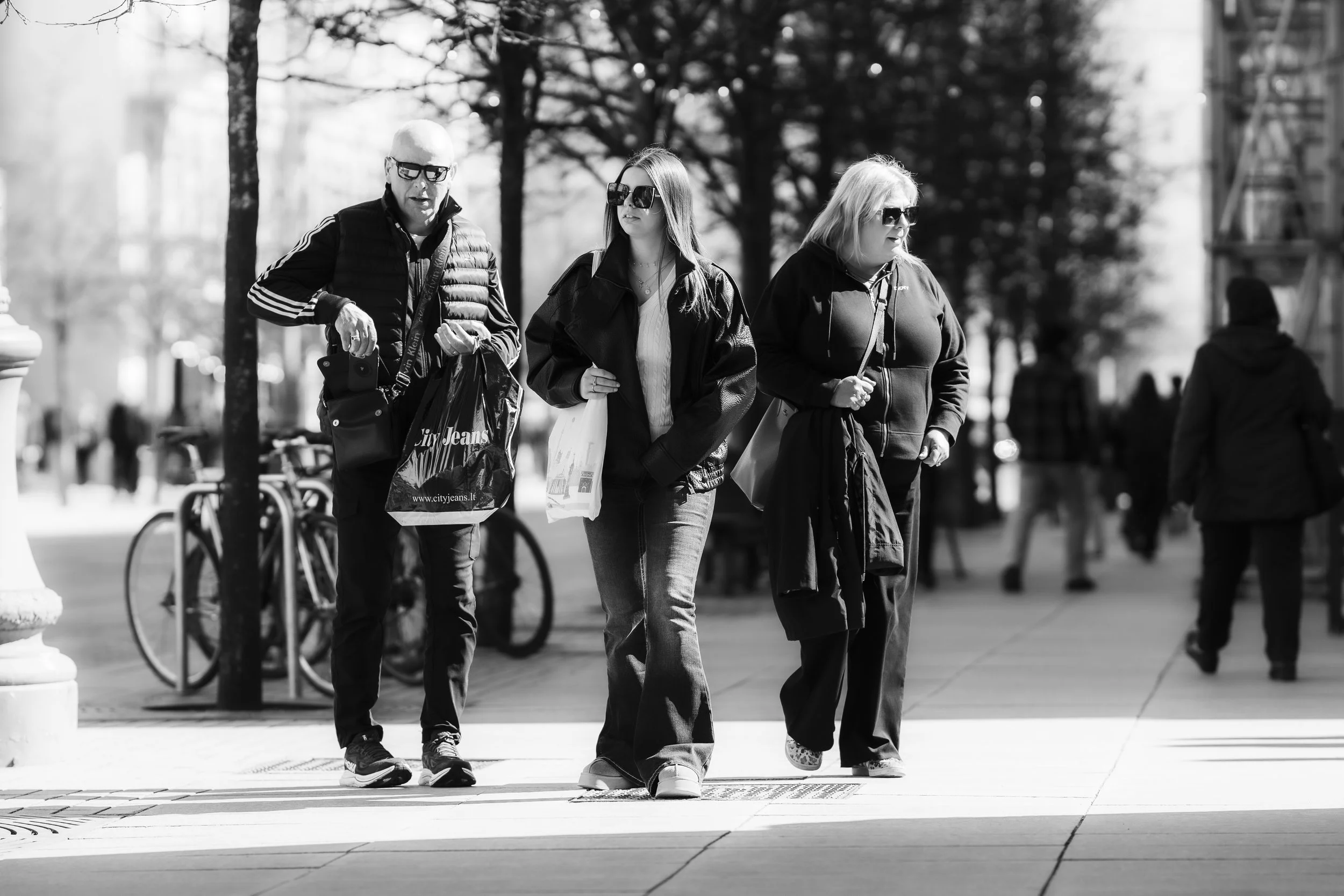 People walking on a city sidewalk, including an older man with glasses and a woman with long hair and sunglasses, all holding shopping bags.