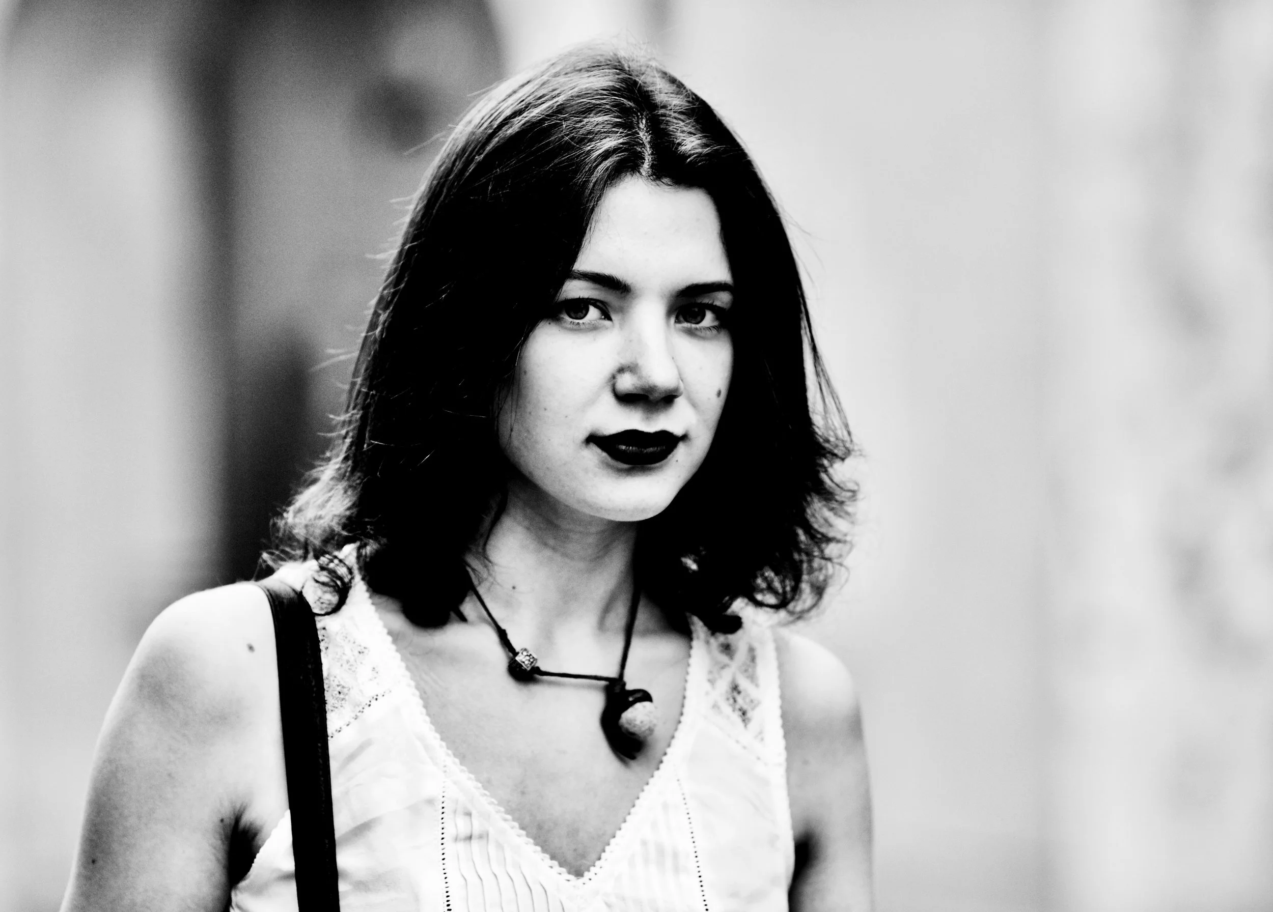 Black and white photograph of a young woman with dark, wavy hair, wearing a light-colored sleeveless top and a necklace with a pendant, walking outdoors.