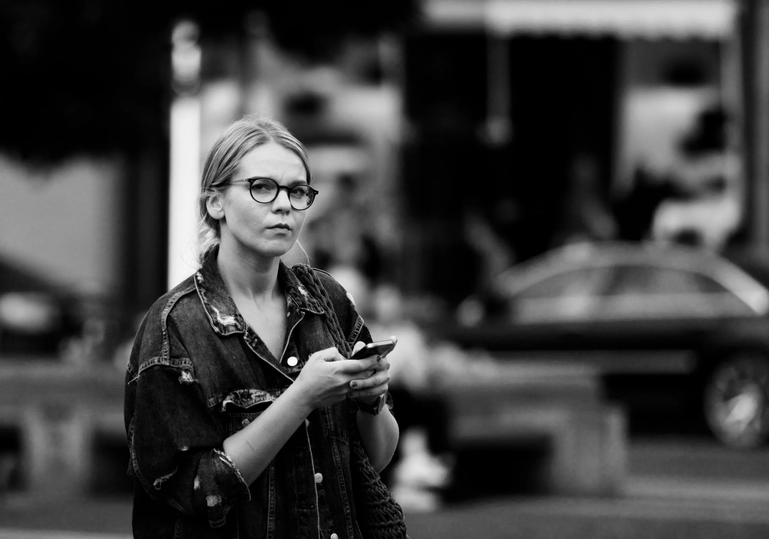 A woman with glasses and long hair is holding a smartphone and looking at the camera. She is wearing a distressed denim jacket. The background shows a blurred street scene in black and white.