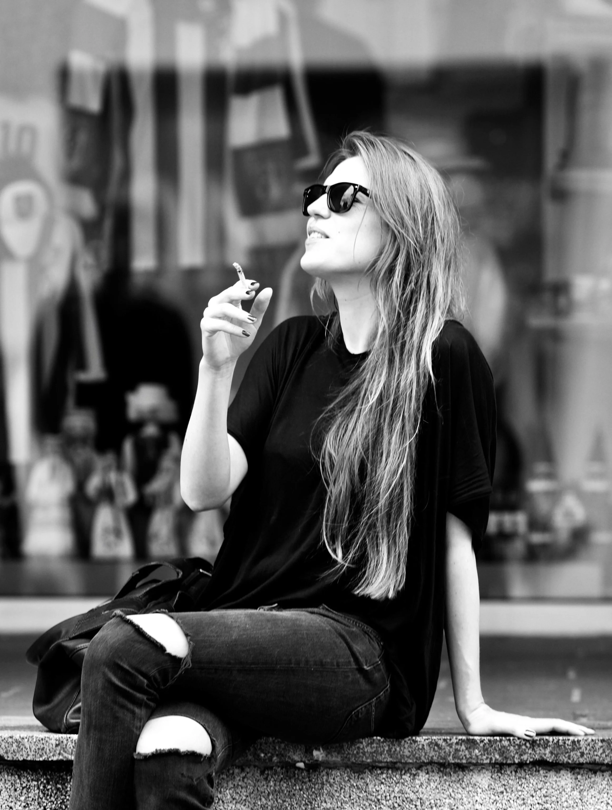 A woman with long hair wearing sunglasses, a black t-shirt, and ripped jeans sitting on a ledge, smiling, while holding a cigarette.