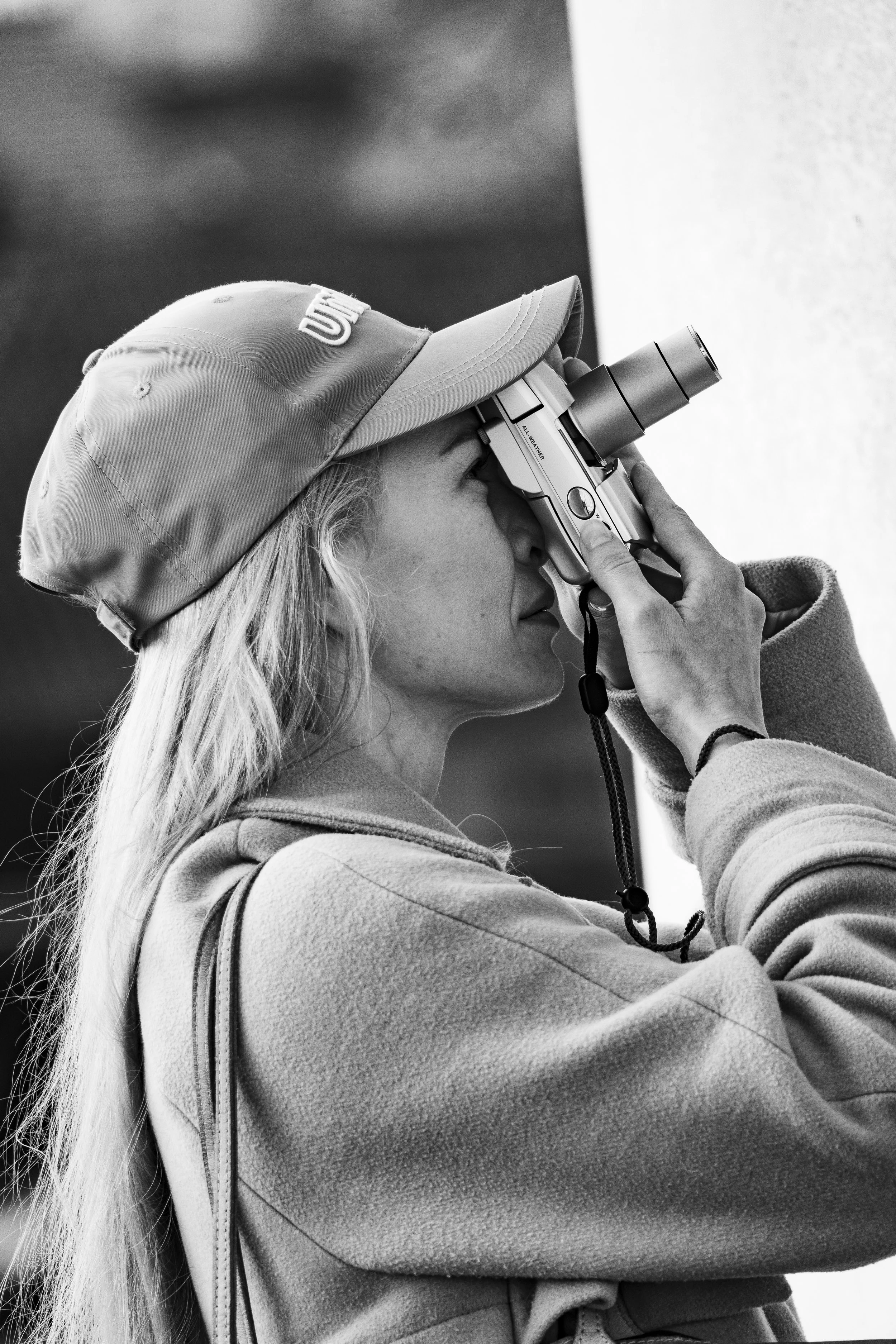 A woman with long hair, wearing a baseball cap and a sweatshirt, looking through a viewfinder of a camera with a large lens, in black and white.
