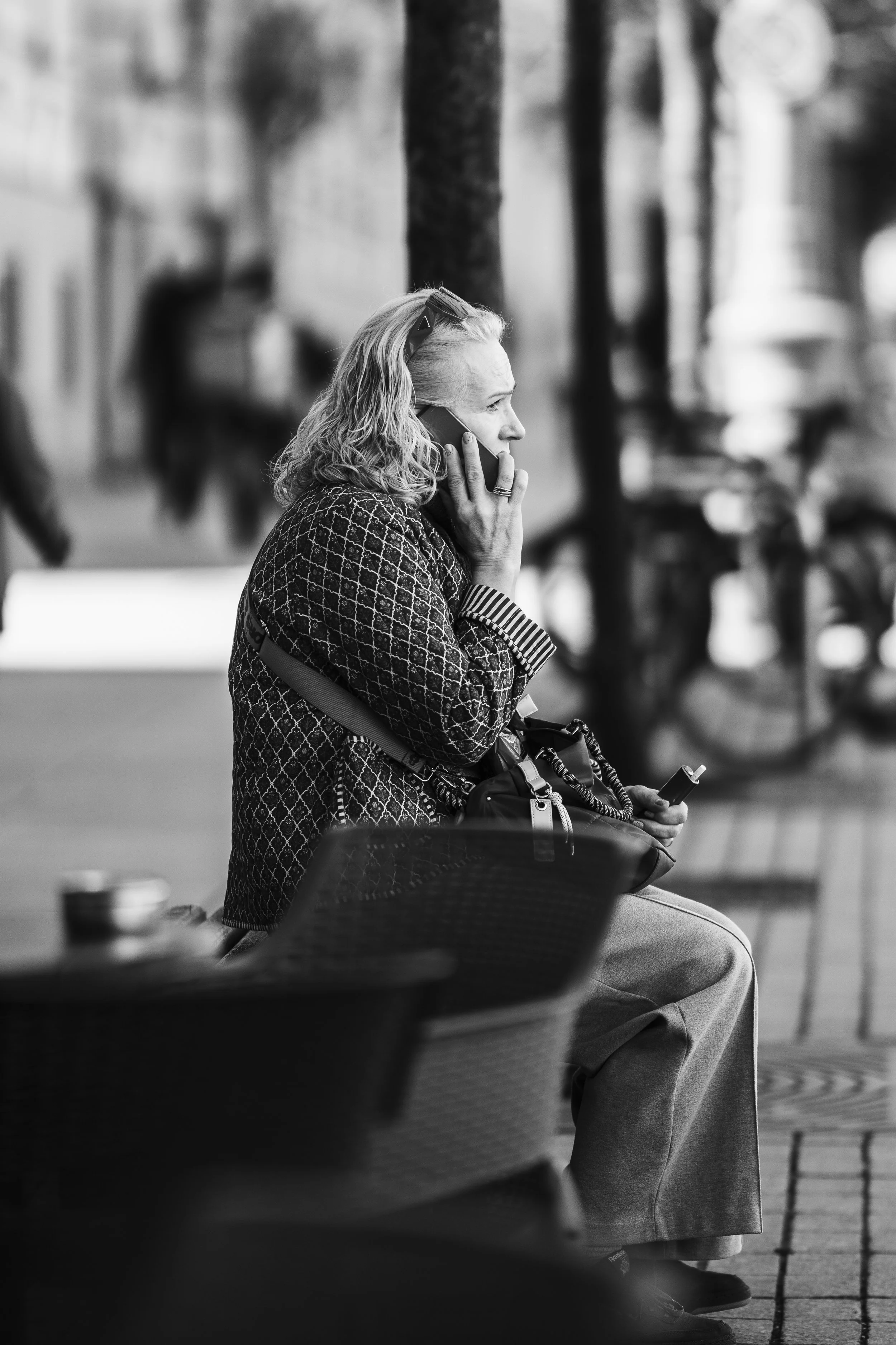 A woman sitting on a bench outdoors, talking on her phone and holding a small object, with trees and bicycles in the background.