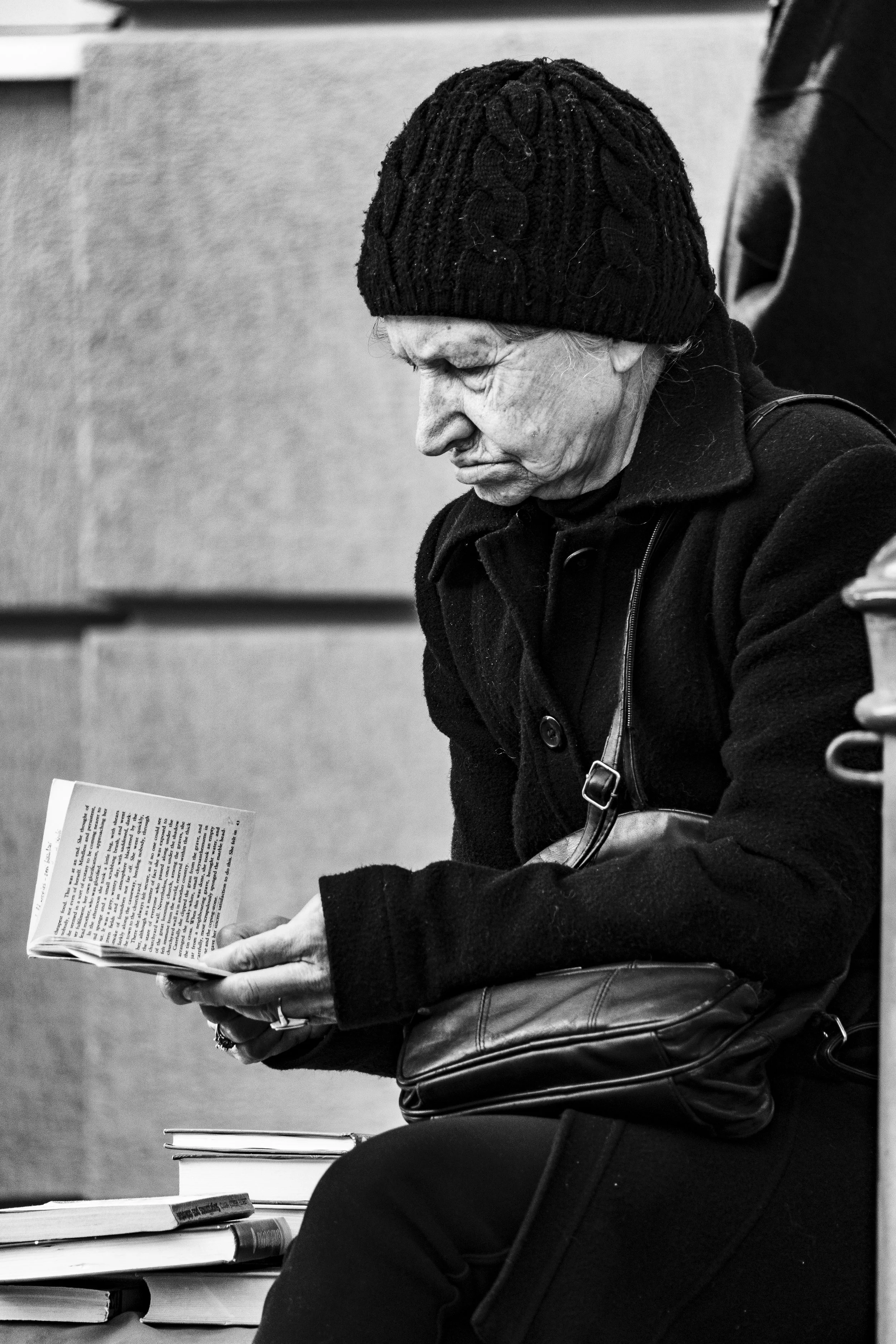 An elderly woman with a knitted hat and dark coat is sitting and reading a book, with a stack of books nearby.