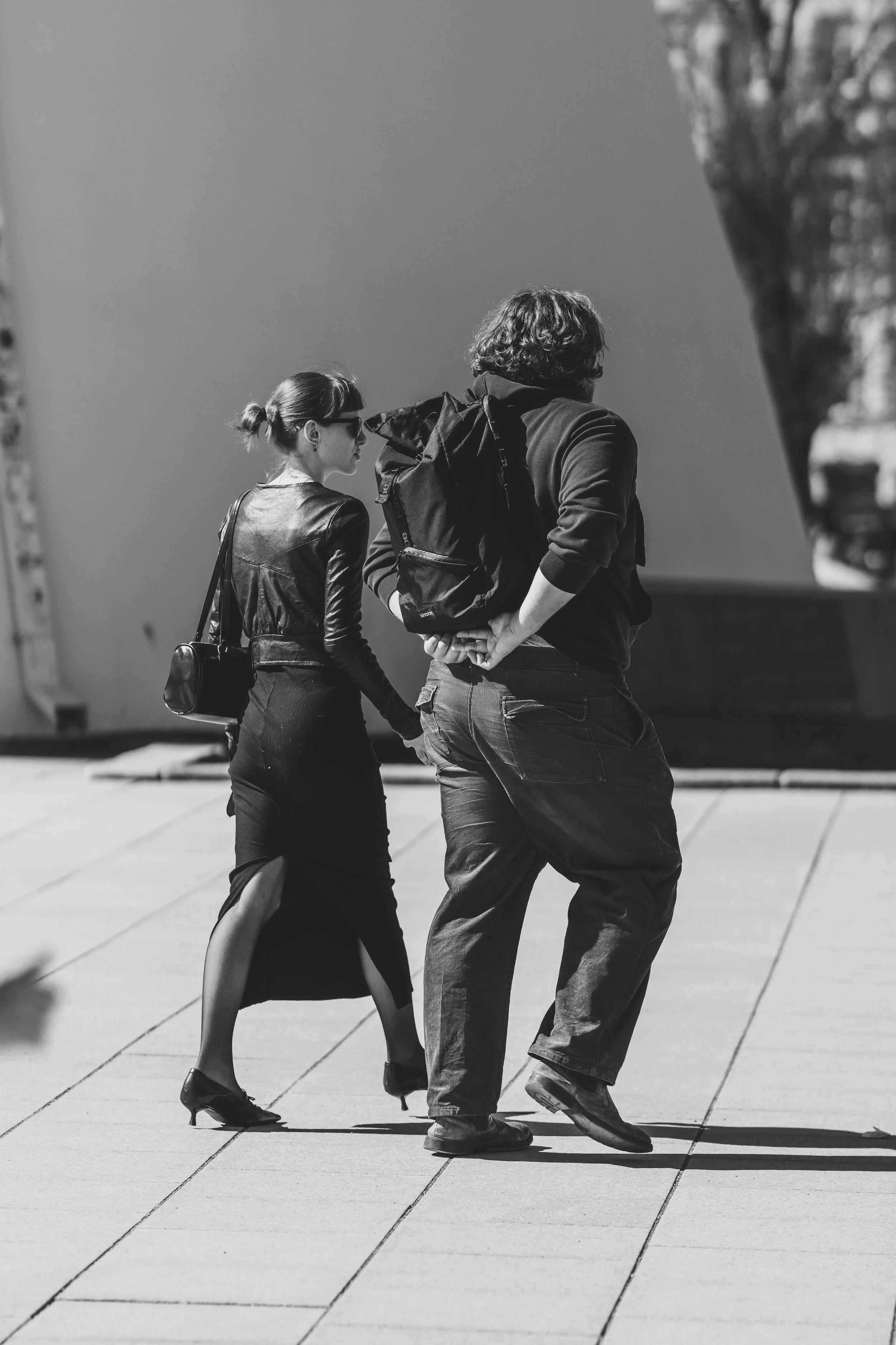 A black and white photo of a woman and a man walking outside on a sunny day. The woman is dressed in a long skirt, high heels, and a leather jacket, carrying a small handbag. The man is dressed casually in jeans and a hoodie, carrying a backpack.
