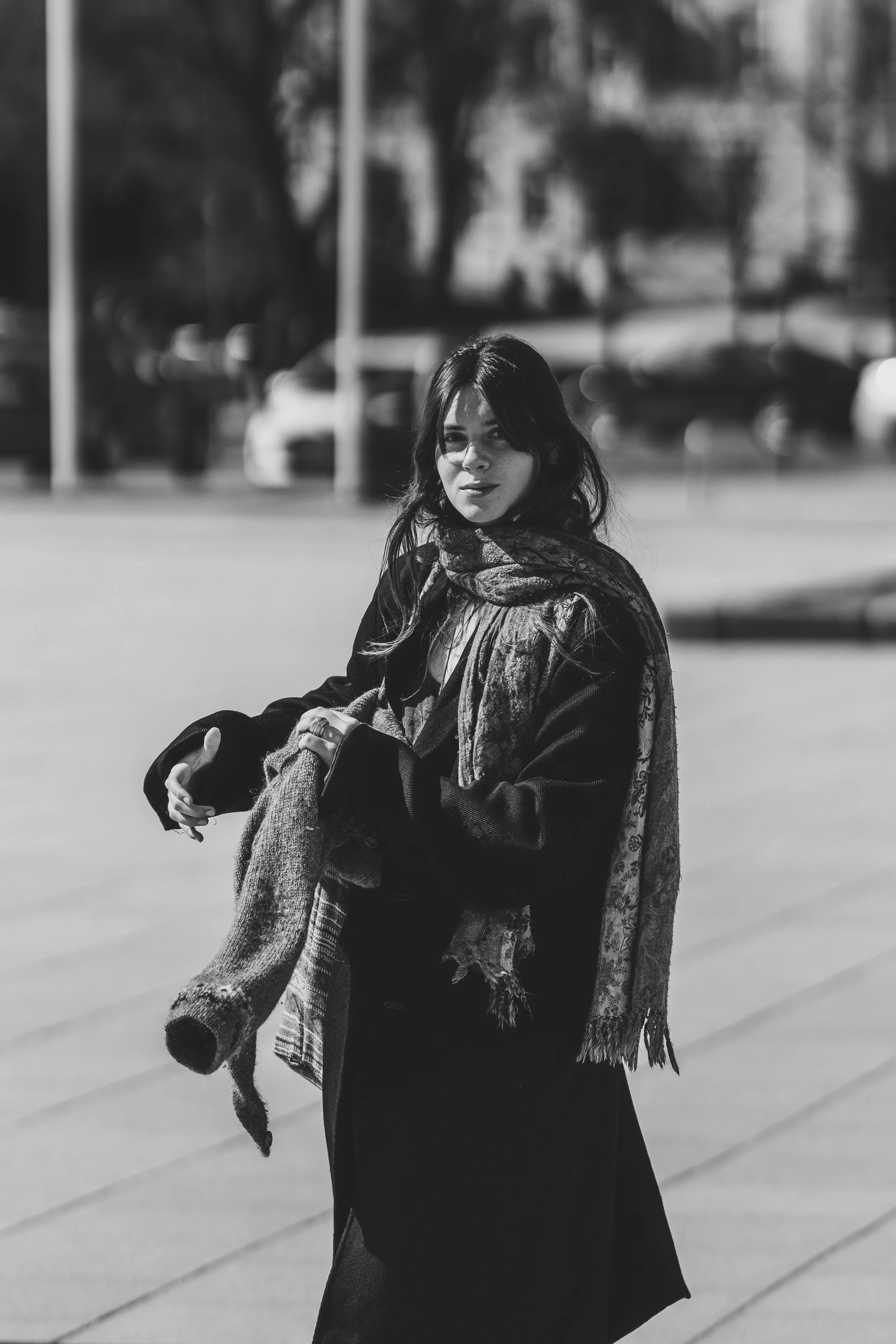A young woman with dark hair and a scarf standing outdoors on a city street at night. She is holding a coat and looking at the camera with a slight smile.