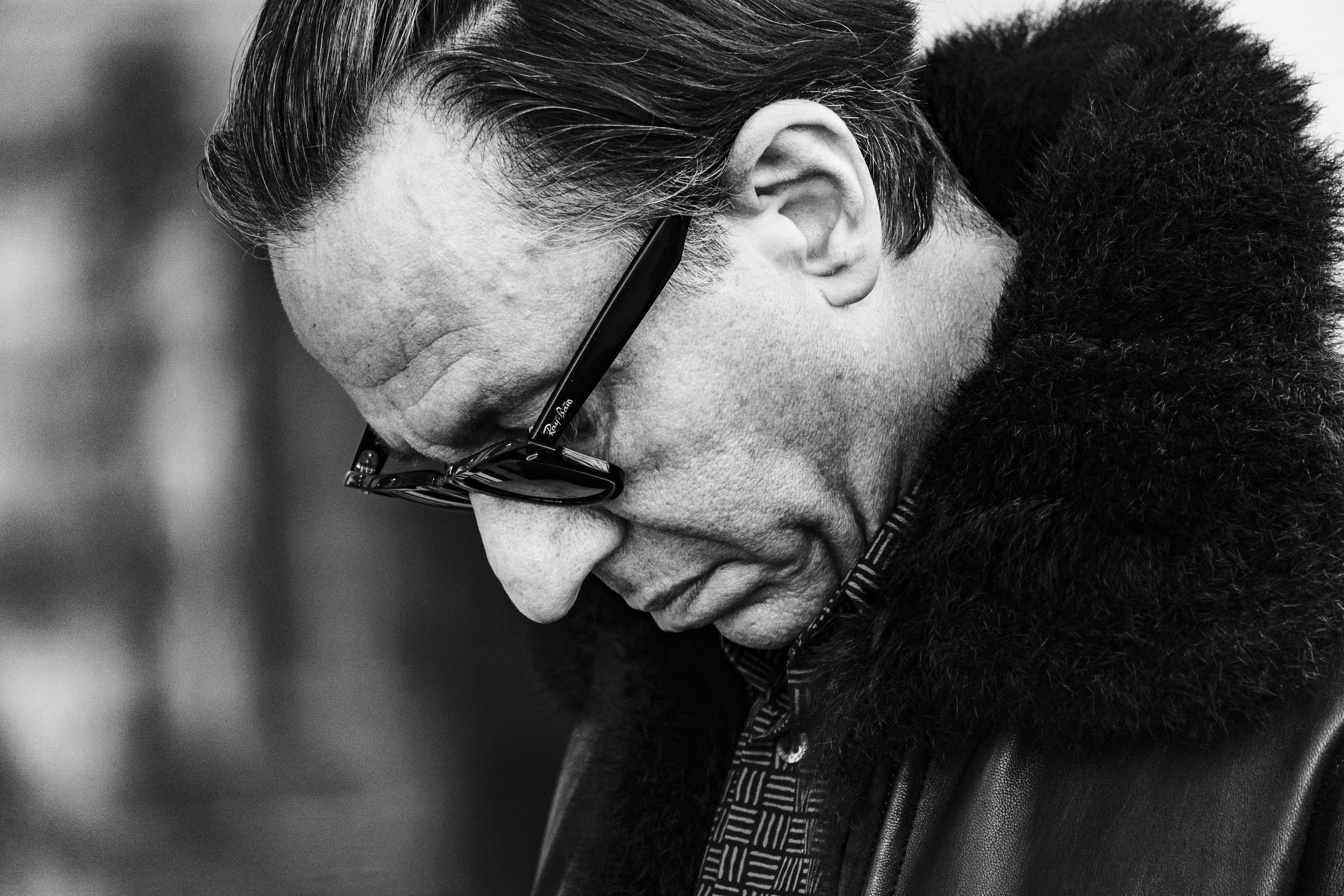 Black and white photo of a man with glasses looking down, wearing a jacket with a fur collar.