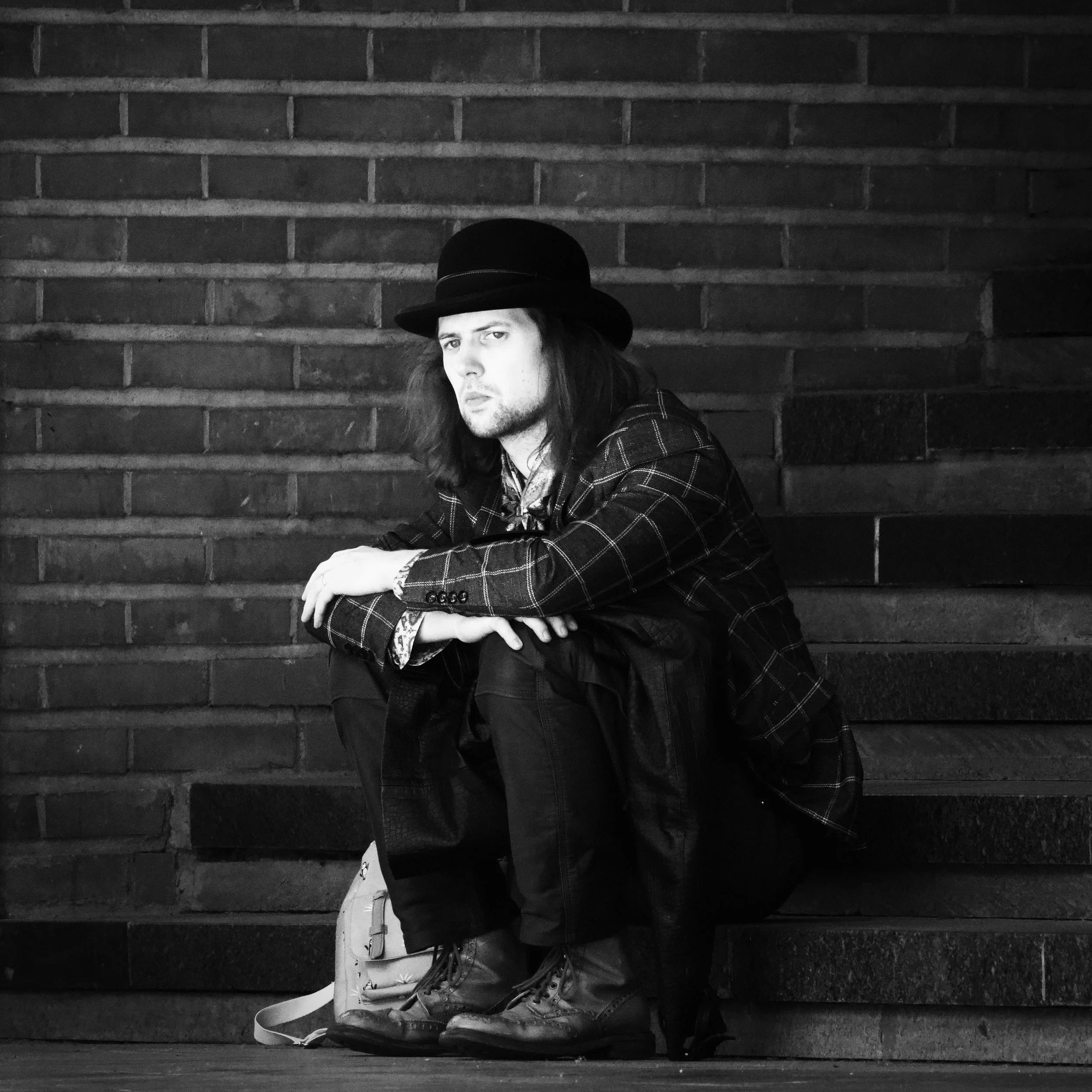 A young man with long hair, wearing a black hat and plaid jacket, sitting on steps against a brick wall, looking at the camera with a serious expression.