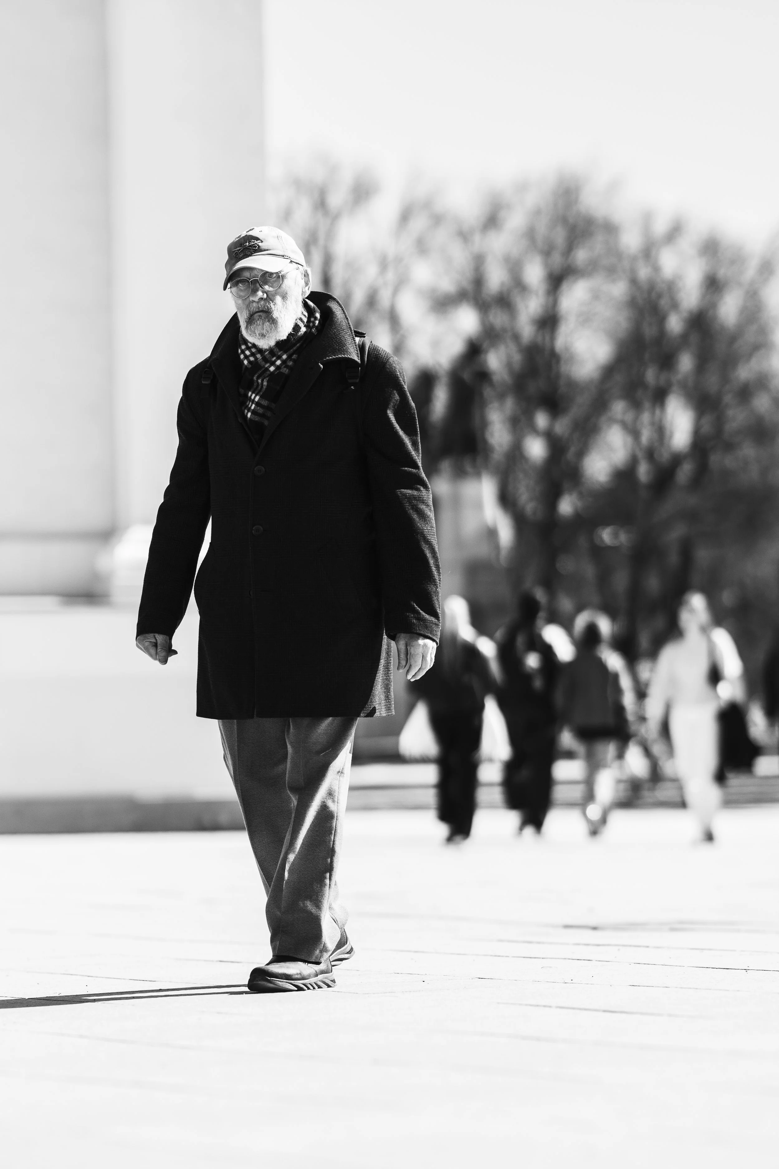 A black and white photo of an elderly man with glasses, a cap, and a coat walking outdoors, with several people and trees in the background.