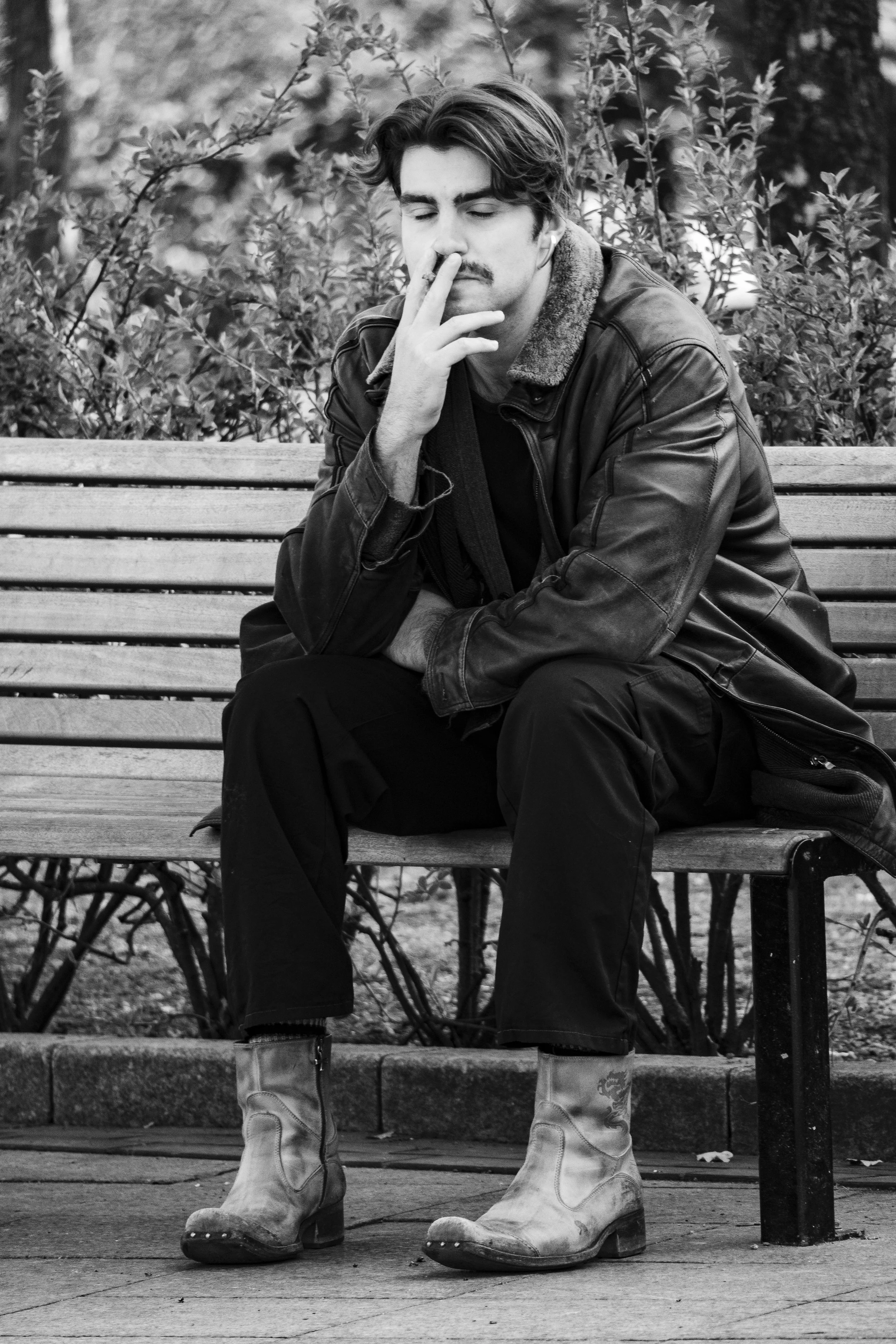 A young man with a mustache sitting on a park bench, wearing a leather jacket, black pants, and cowboy boots, with eyes closed and fingers near his nose, in a black and white photo.