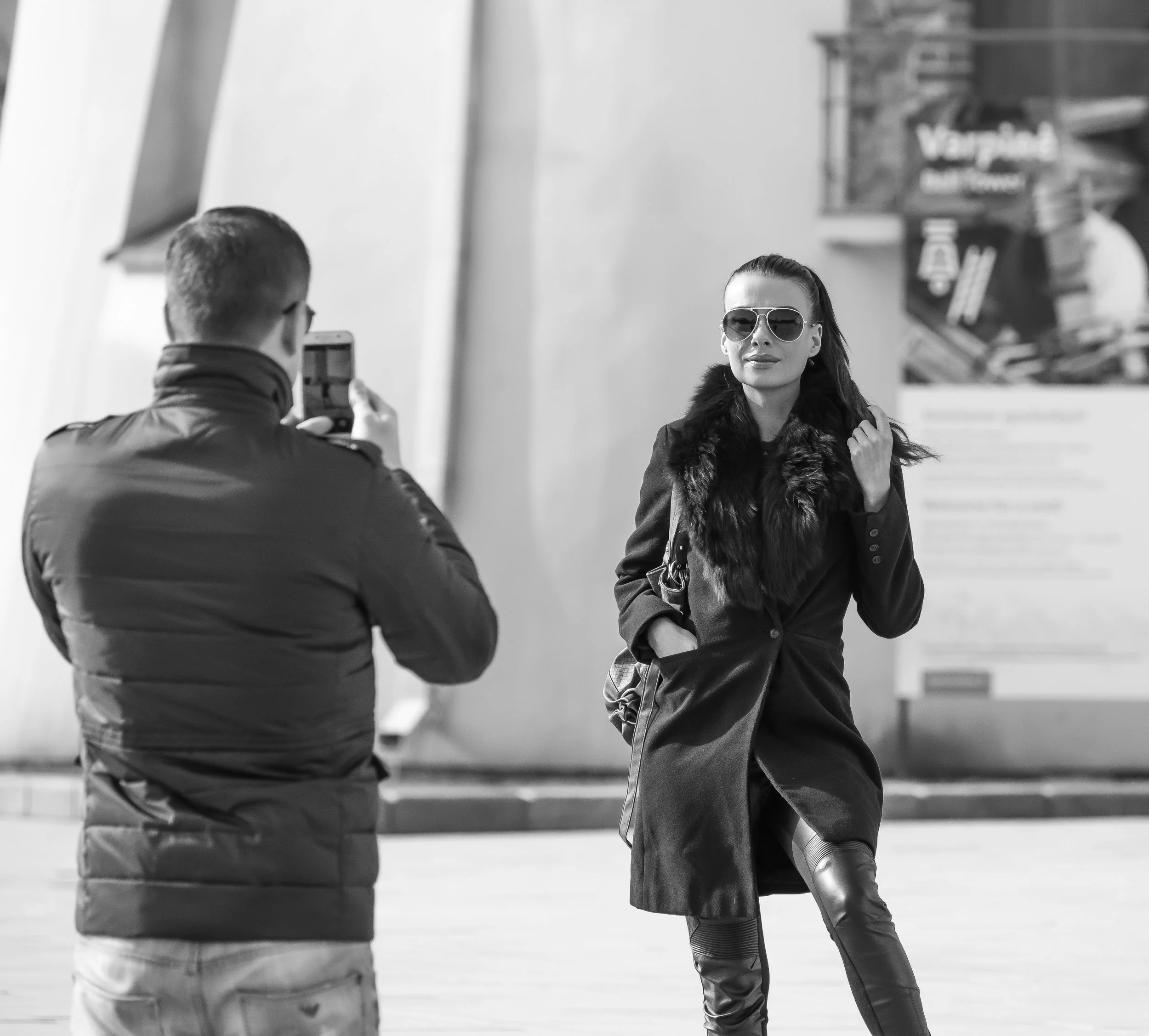 A woman wearing sunglasses and a stylish coat with a fur collar poses for a photo being taken by a man holding a smartphone outdoors.