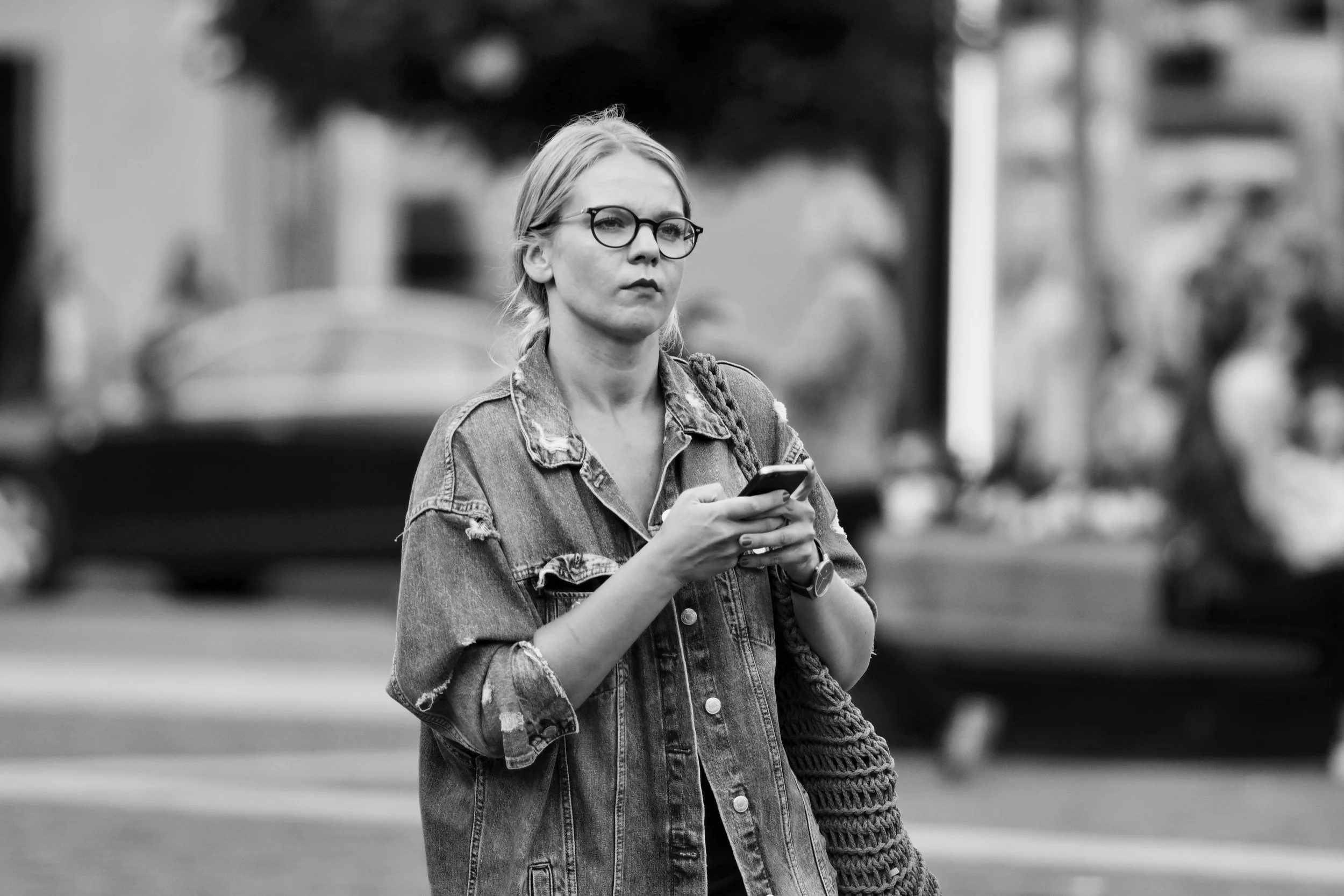 Black and white photo of a woman with glasses and blonde hair holding a phone, standing outdoors in an urban setting.