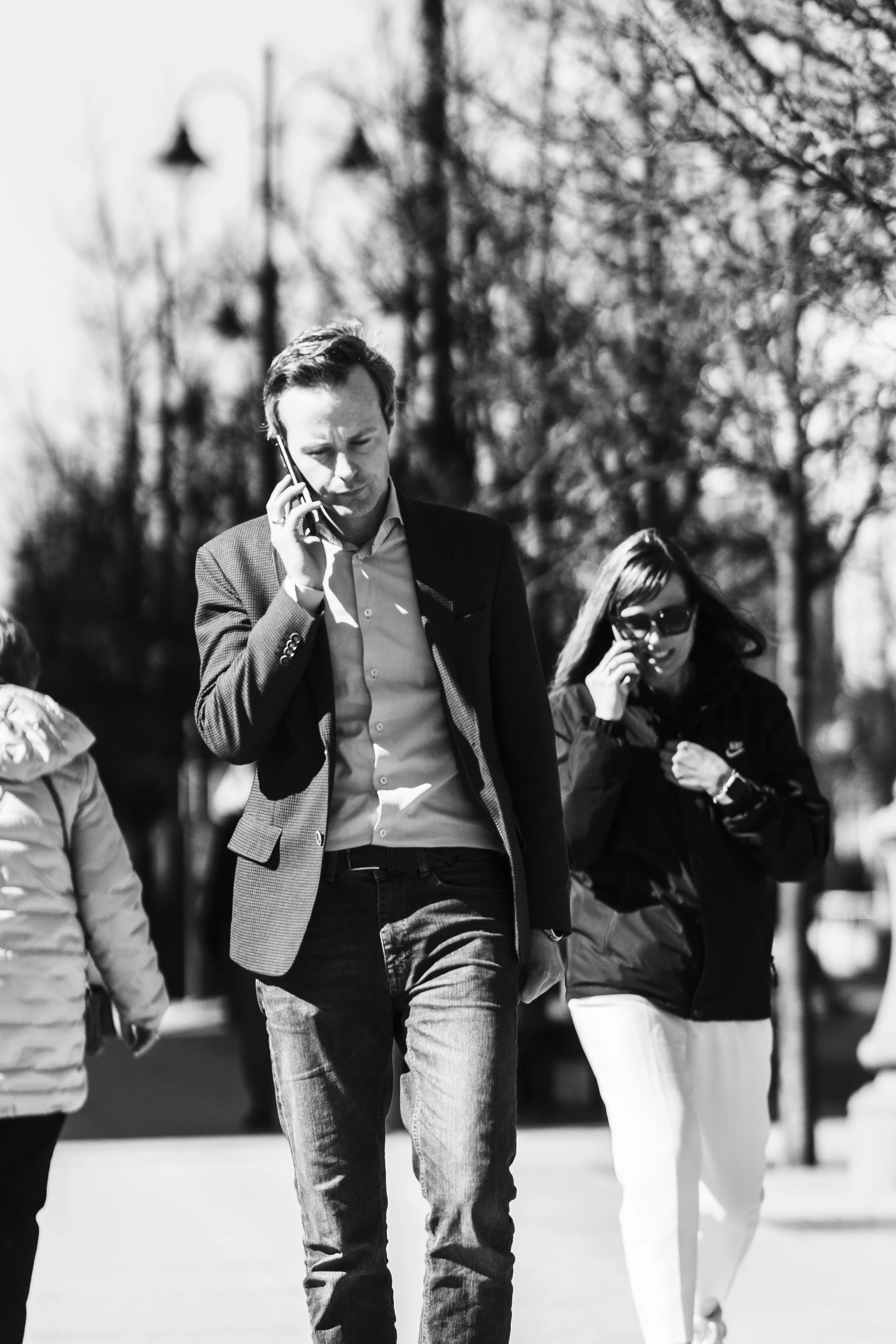 A black and white photo showing a man and woman walking outdoors, both talking on cell phones. The man is wearing a blazer and jeans, and the woman is wearing sunglasses, a jacket, and white pants. The background features trees and street lamps.